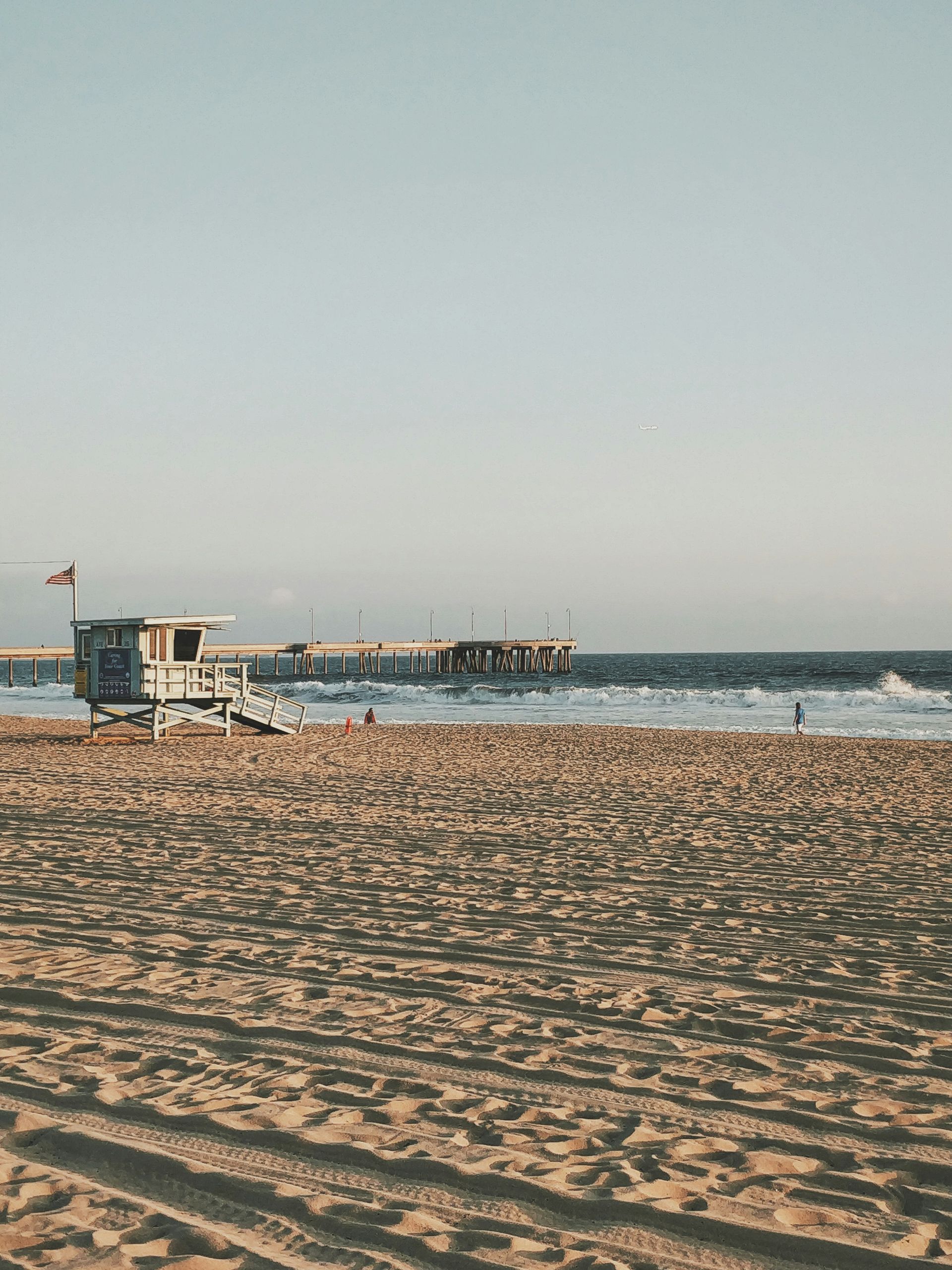 Lifeguard stand on sandy beach with a pier in the distance; blue ocean and sky.