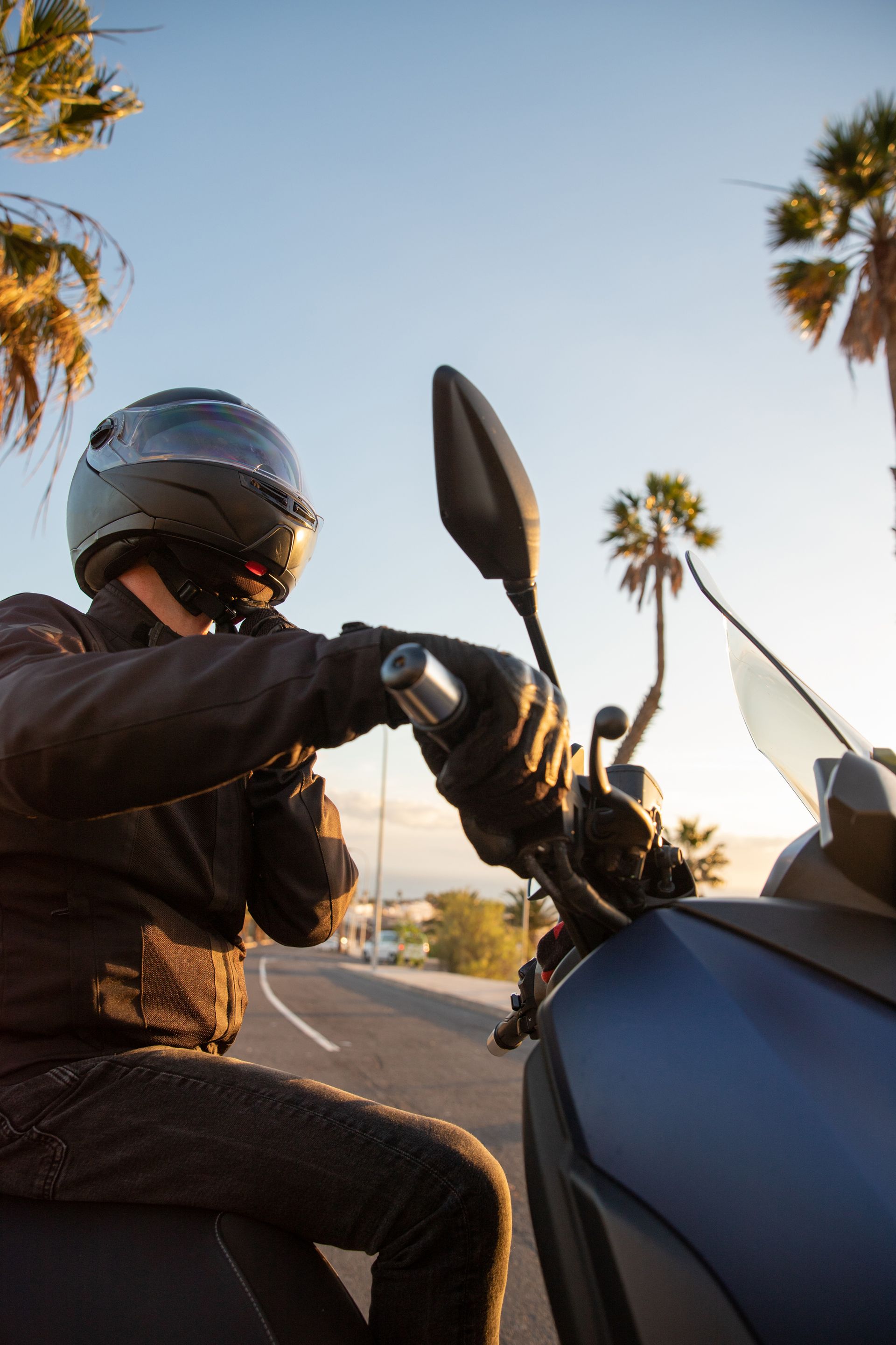 Person on scooter wearing a helmet and gloves, riding on a road with palm trees at sunset.