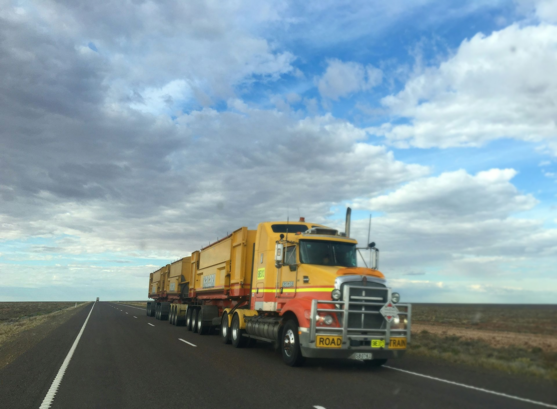 Yellow and red road train truck driving on an asphalt highway under a cloudy sky.