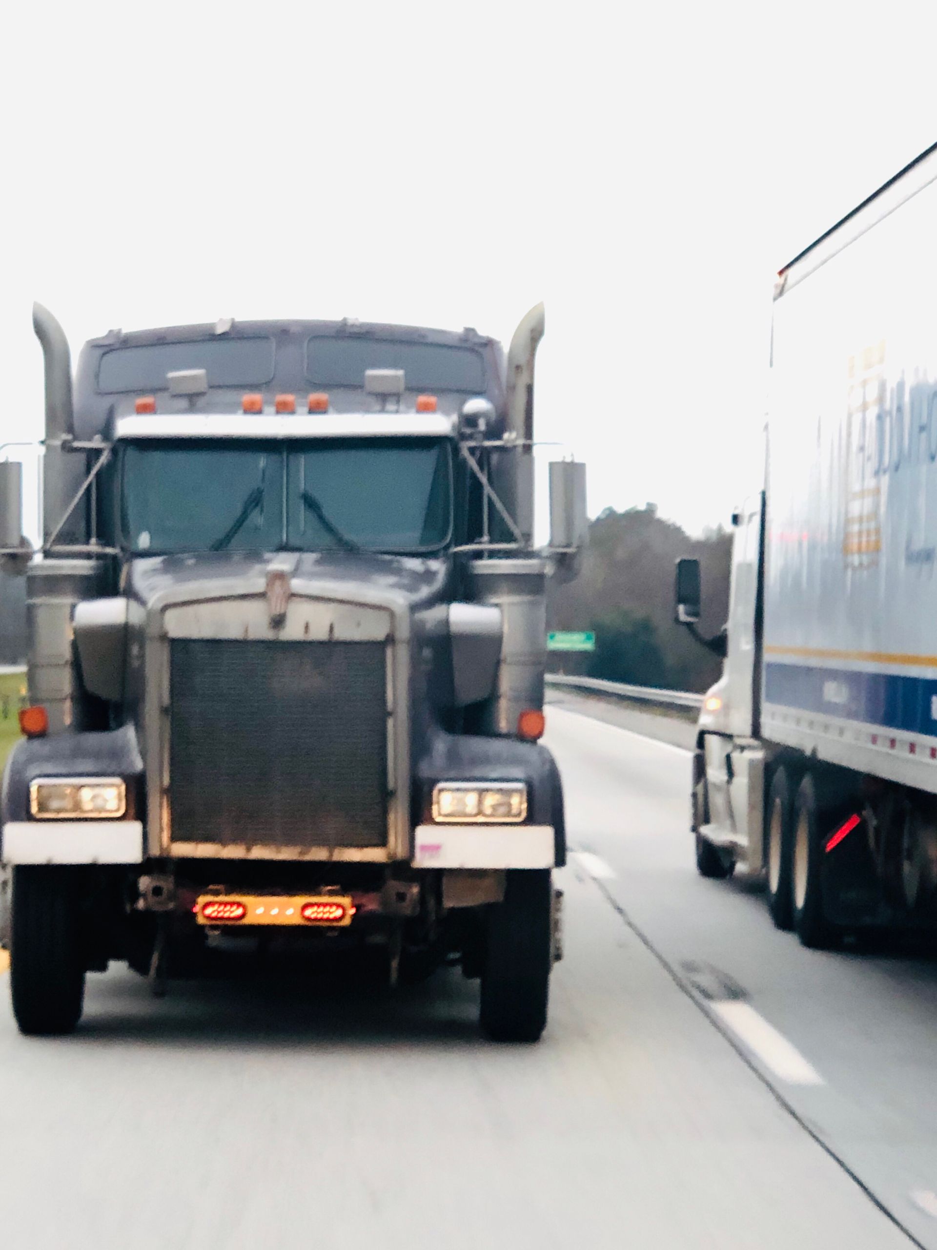 Dark gray semi-truck driving on a highway, another truck to the right. Overcast skies.