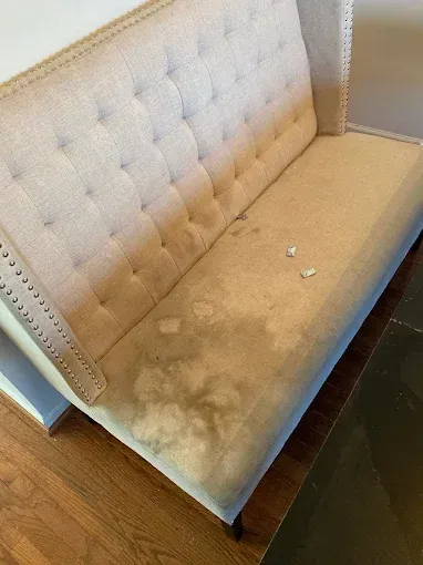 Beige upholstered bench with dark stains on the seat and back, featuring button tufting and nailhead trim.