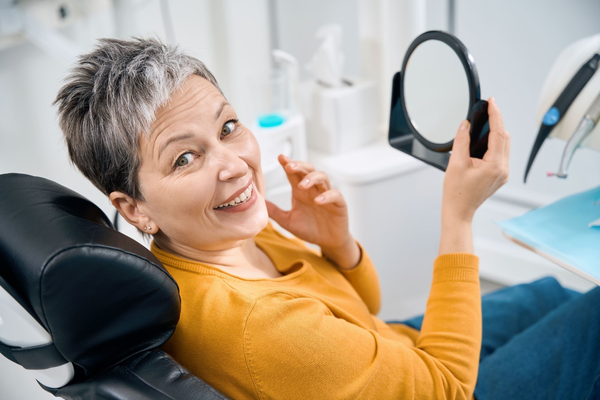Woman sitting in a dental chair holding a mirror and smiling while looking at her teeth after dental crown restoration treatment.