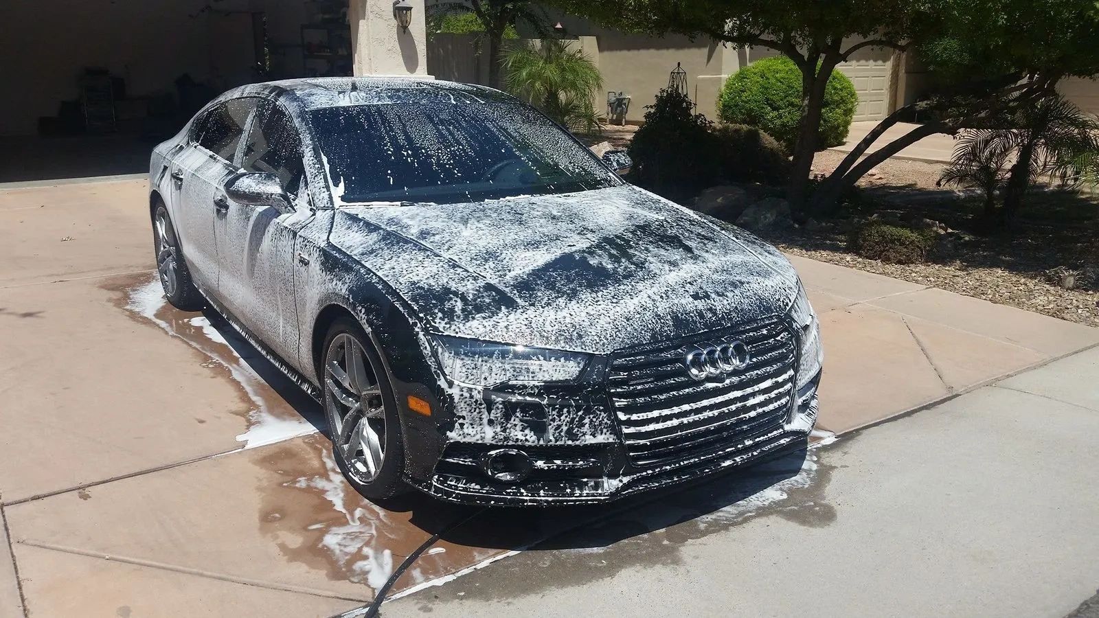 White sedan covered in soap suds being washed in a driveway on a sunny day