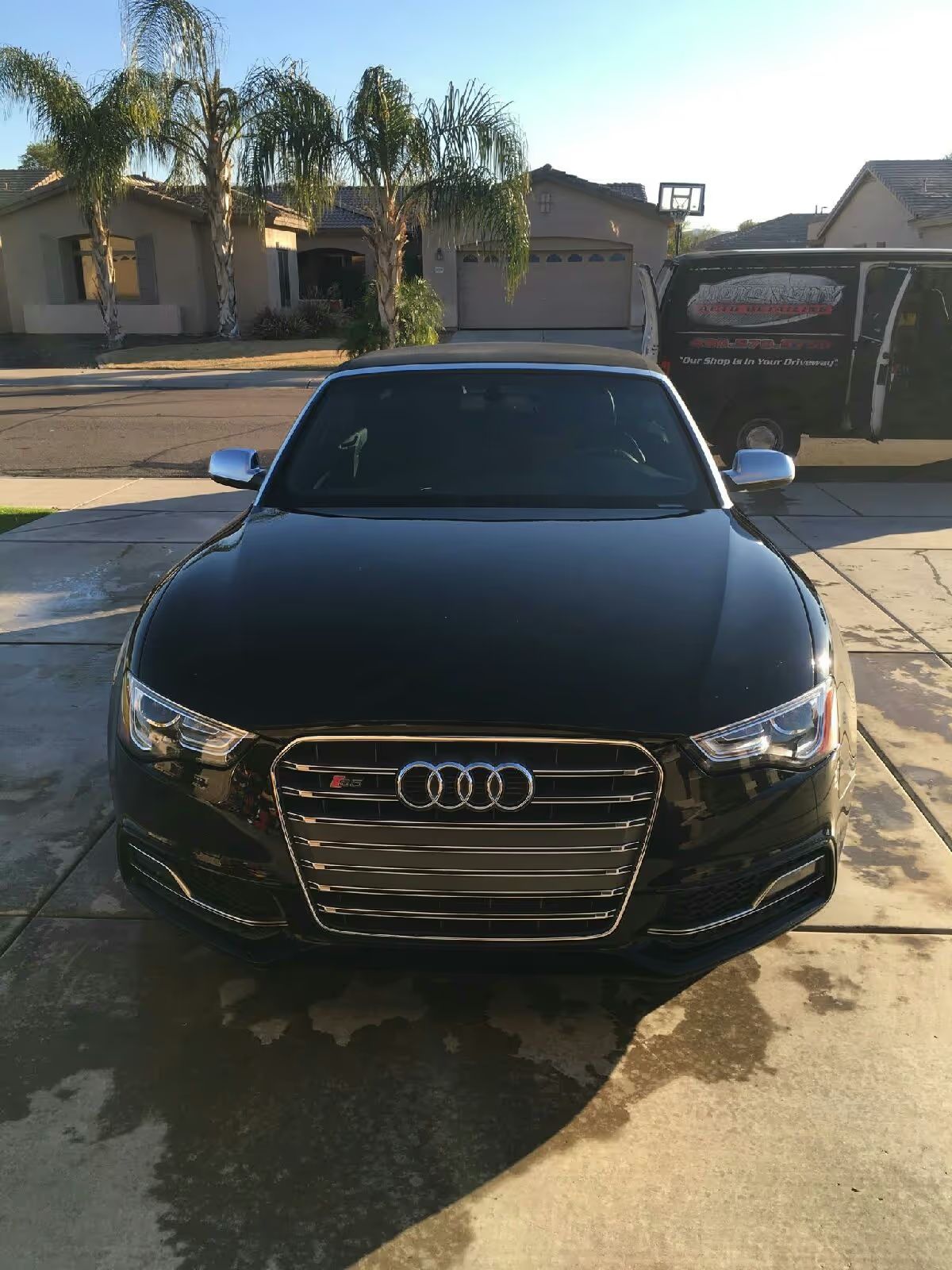 Black Audi sedan parked in a driveway in front of a house, with sunlight and palm trees in the background