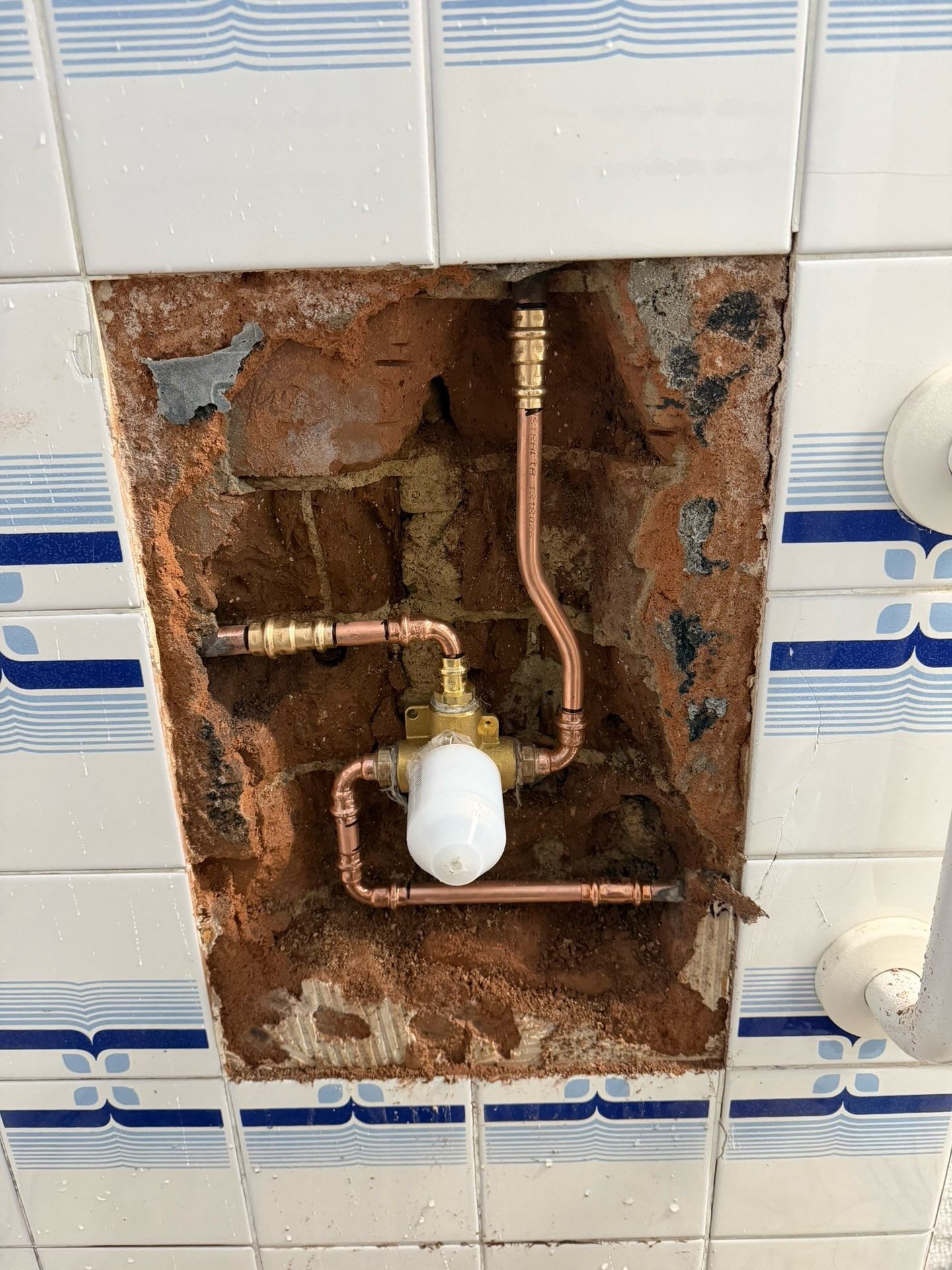 Exposed copper pipes and a wall valve inside a damaged recessed utility box on a tiled wall