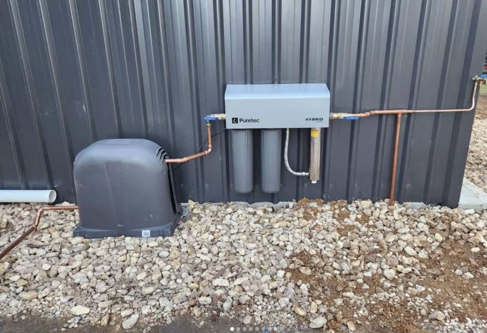 Outdoor utility equipment mounted on a corrugated wall with pipes and a gray cover, above gravel ground