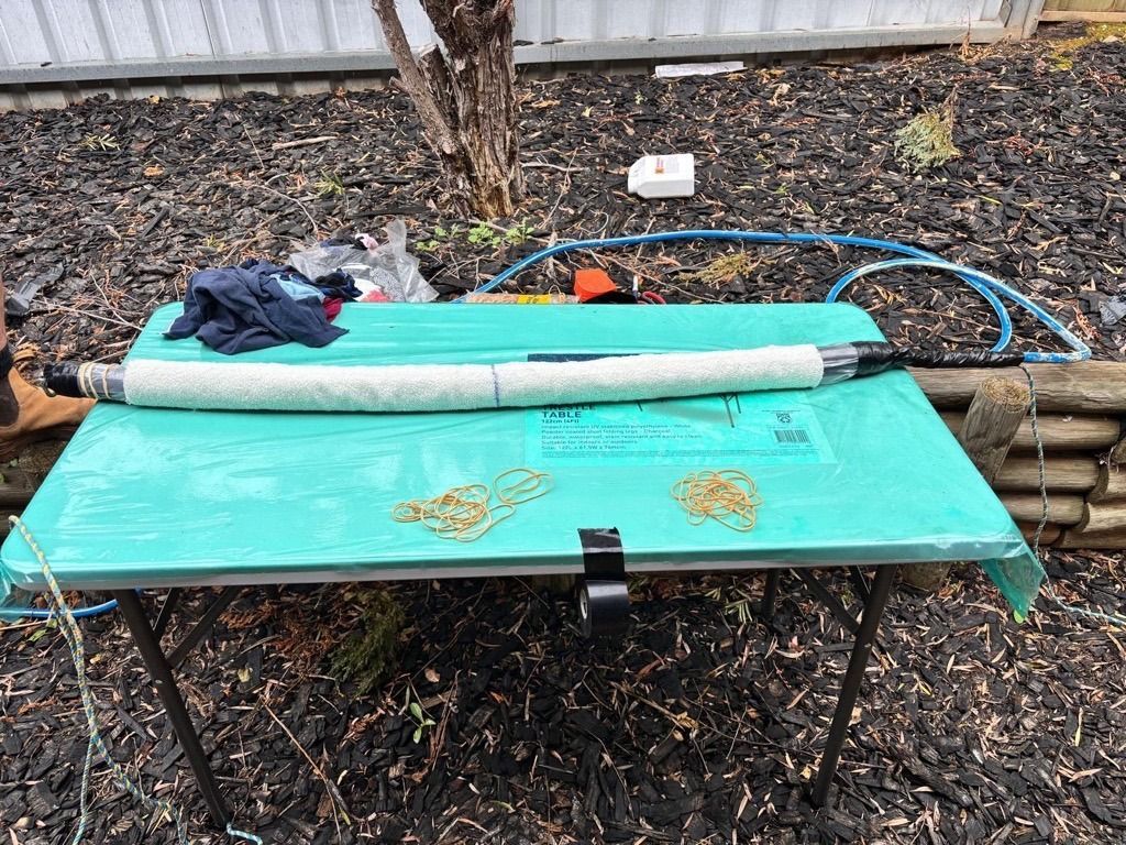 Black folding table outdoors with a turquoise mat, rolled foam tubes, gloves, and a blue hose on mulch