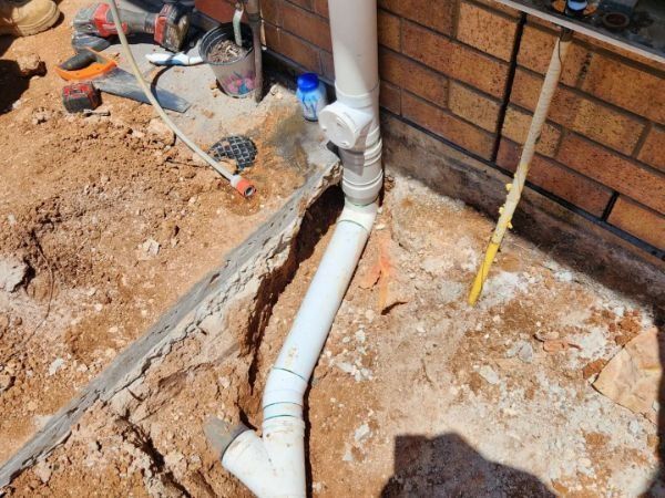 White PVC drain pipe installed in a dirt trench beside a brick house wall.