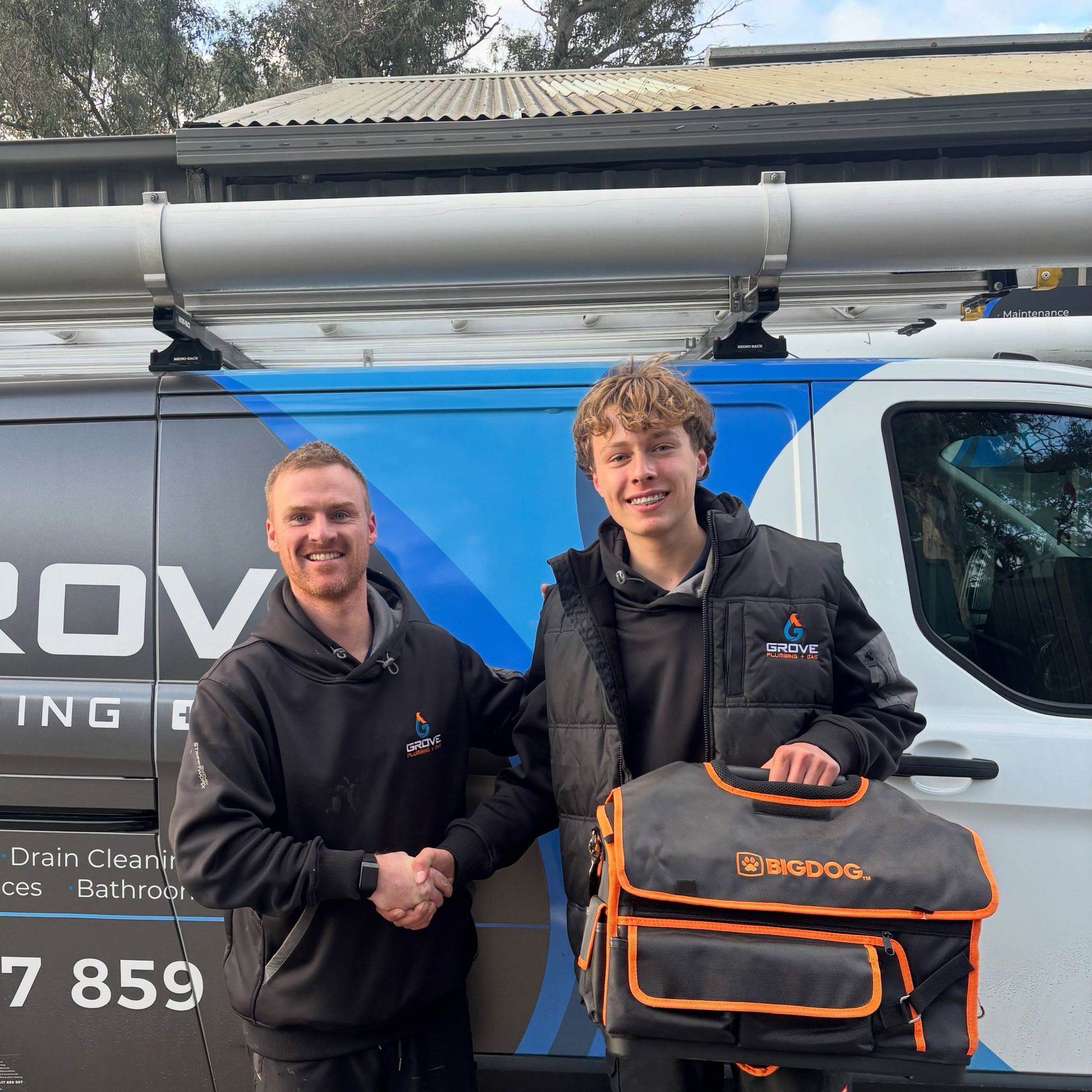 Man standing beside a Grove Plumbing & Gas service van with blue graphics and tools on the roof rack