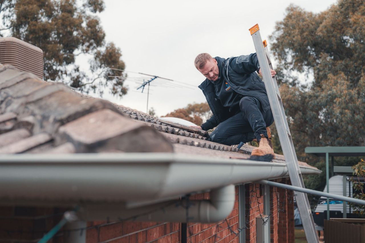 Worker repairing a rooftop gutter beside a brick house, with a ladder and trees in the background
