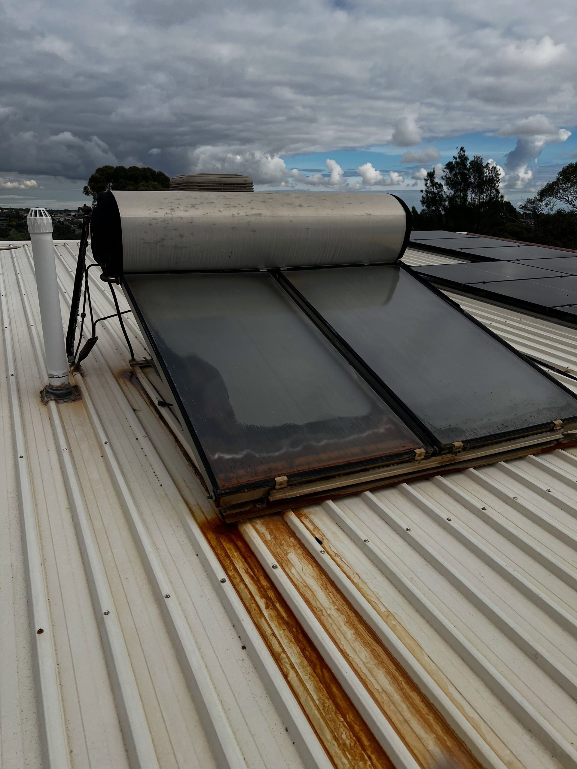Rooftop solar water heater with black panels and cylindrical tank on a corrugated metal roof under cloudy sky