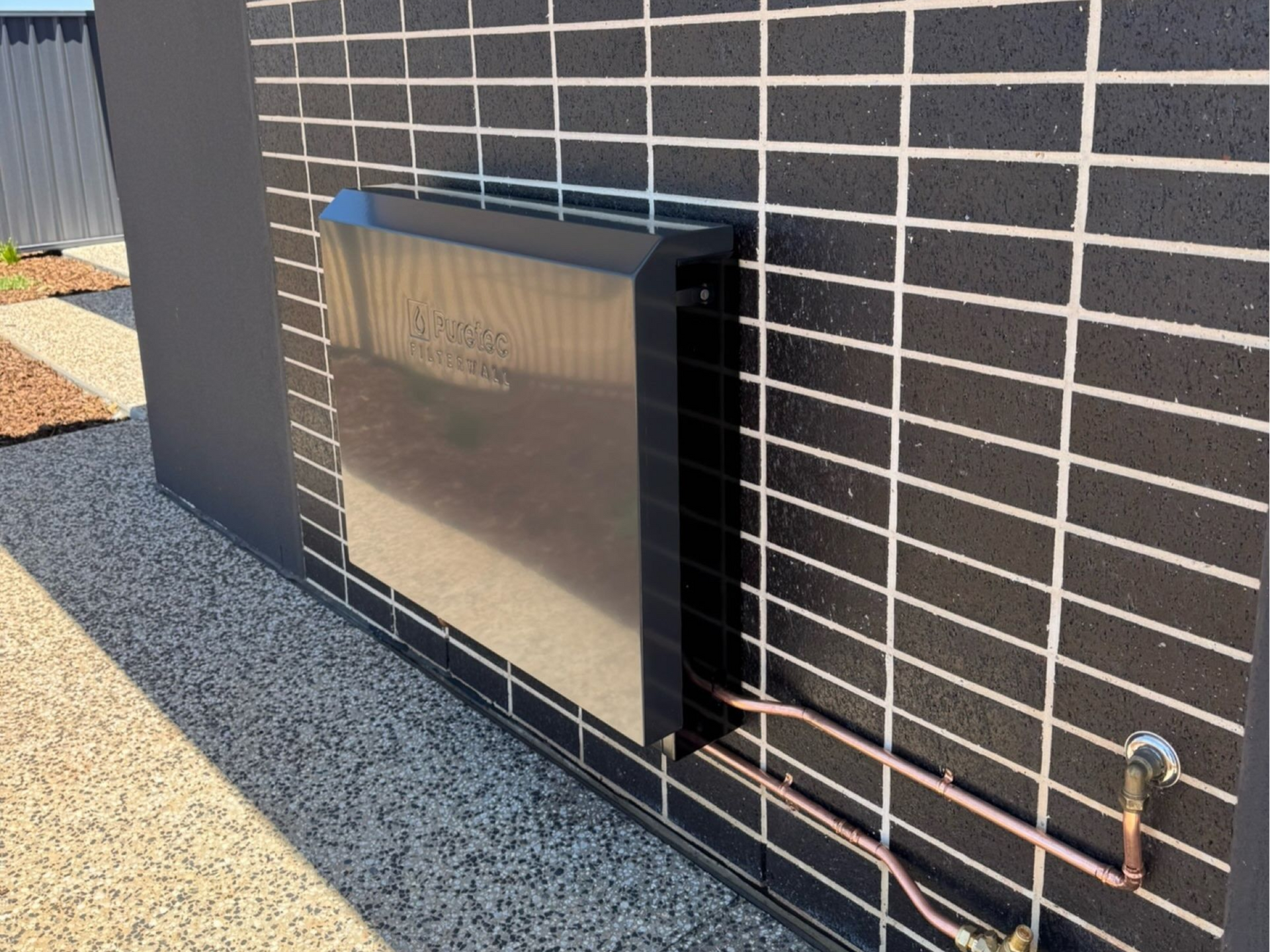 Black wall-mounted utility box on a brick wall with exposed copper pipes running along the base.