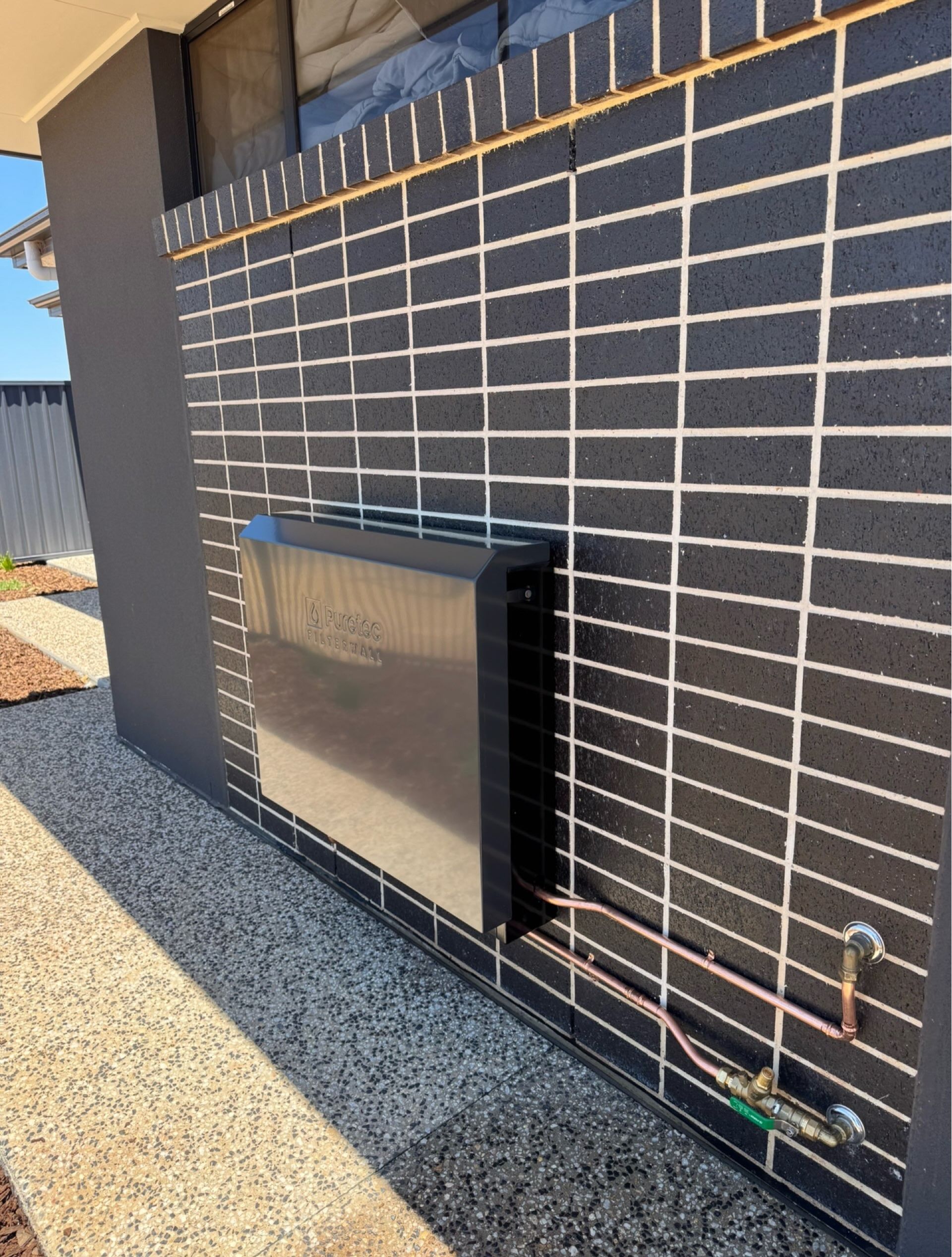 Exterior wall with black brick cladding, a mounted metal unit, and exposed pipes on gravel ground