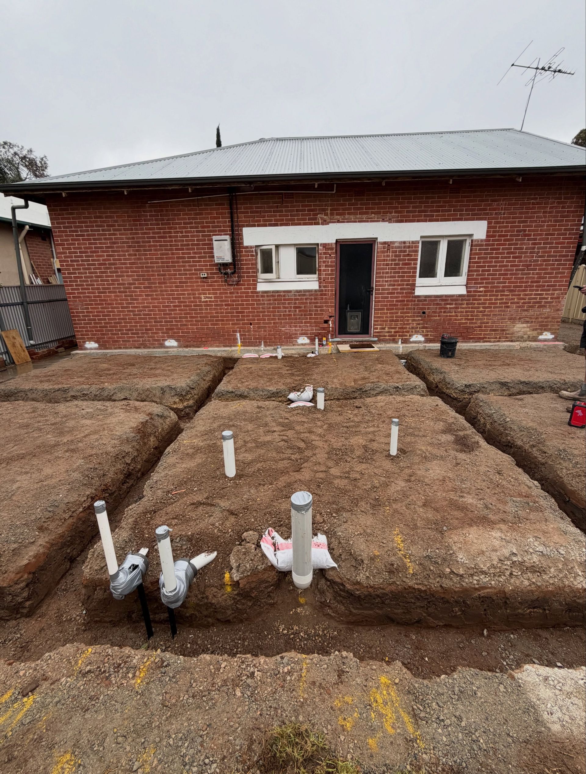 Small brick house with a metal roof and a rough garden plot marked by white pipes in the foreground.