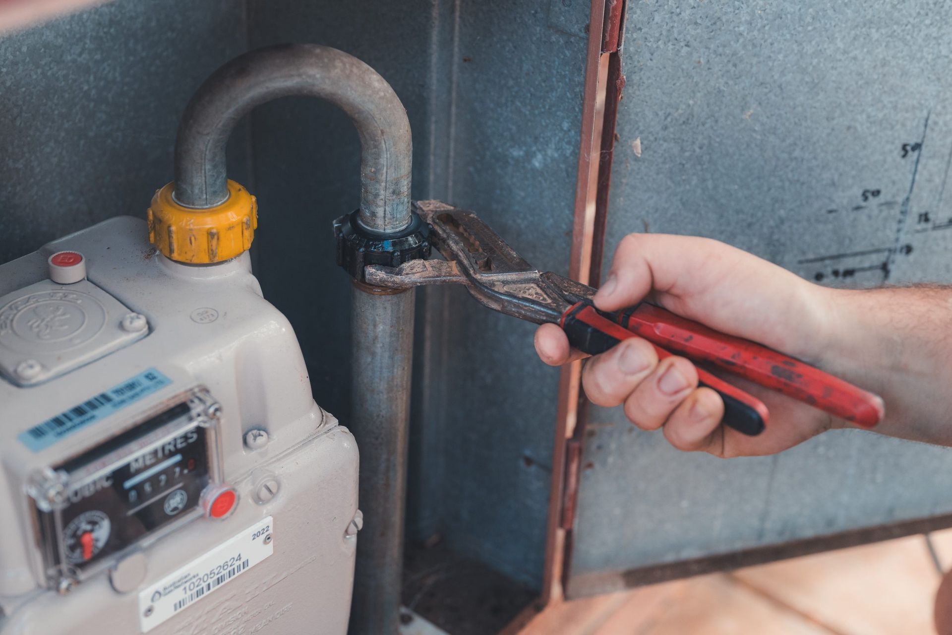 Hand holding pliers tightening a pipe joint next to a gas meter and metal piping