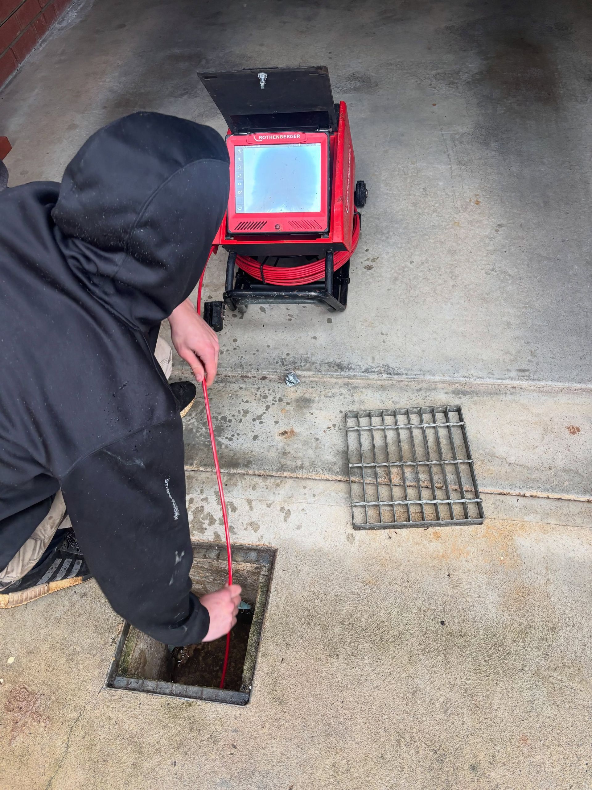 Worker in black hoodie inspecting a sewer drain with a red inspection camera on wet concrete.