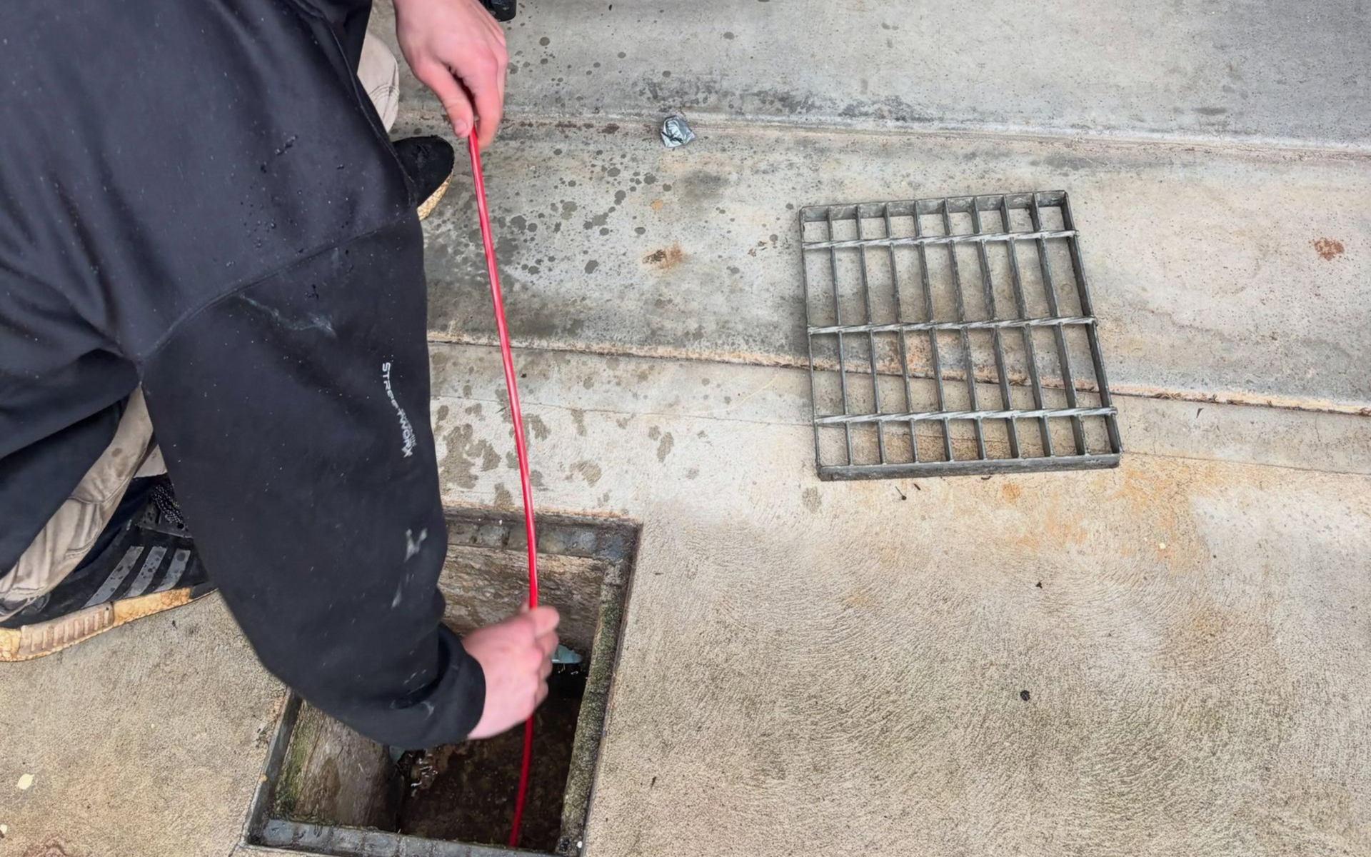 Person lowering a red measuring tape into a floor drain beside a metal grate on a concrete floor