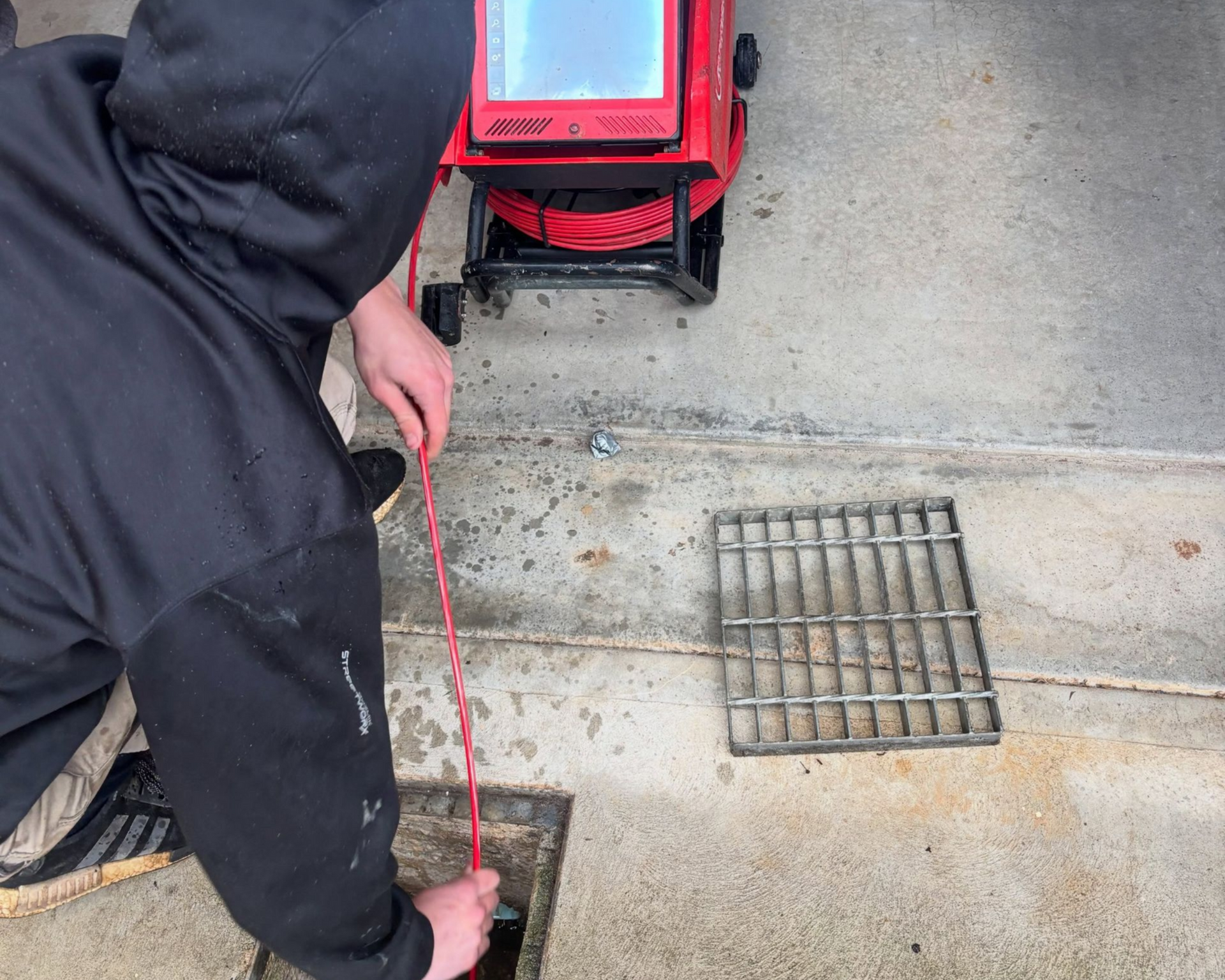 Person using a red cable near a drain grate on a concrete floor beside a red machine.