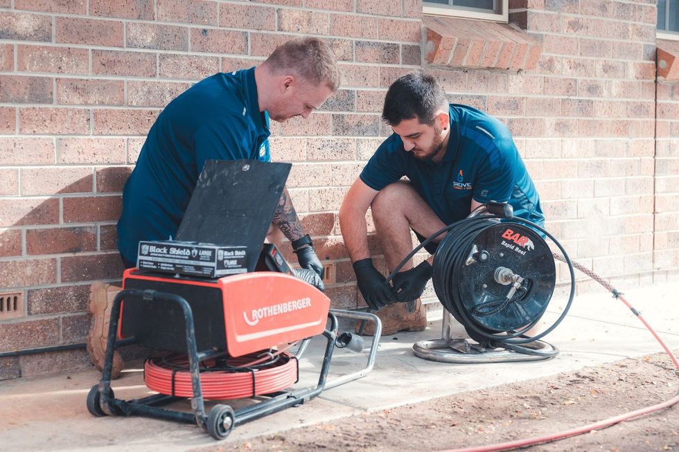 Two workers crouch beside a red welder and fan on a brick patio, repairing or setting up equipment.