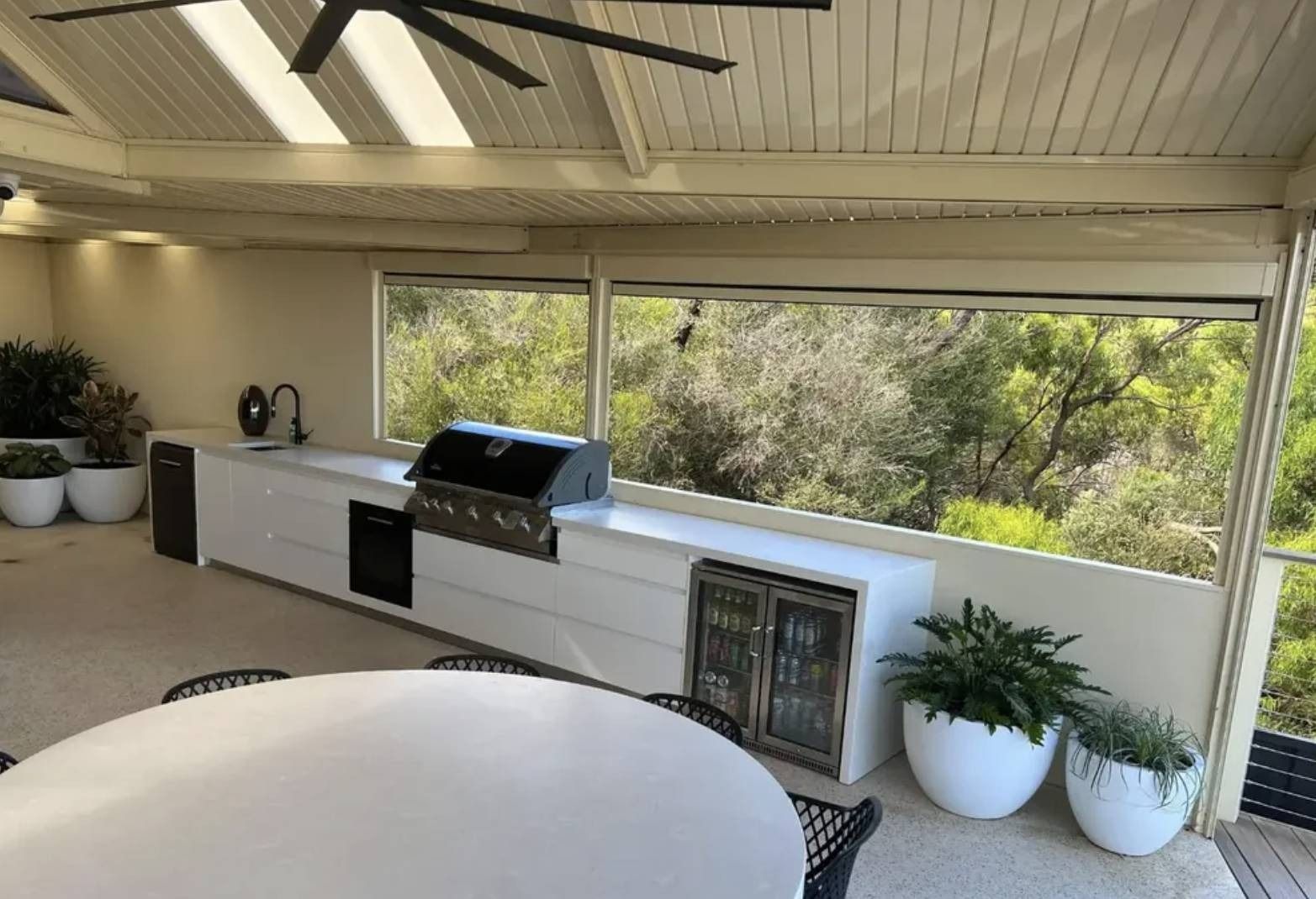 Covered patio with built-in grill, white cabinets, potted plants, and a view of greenery beyond.