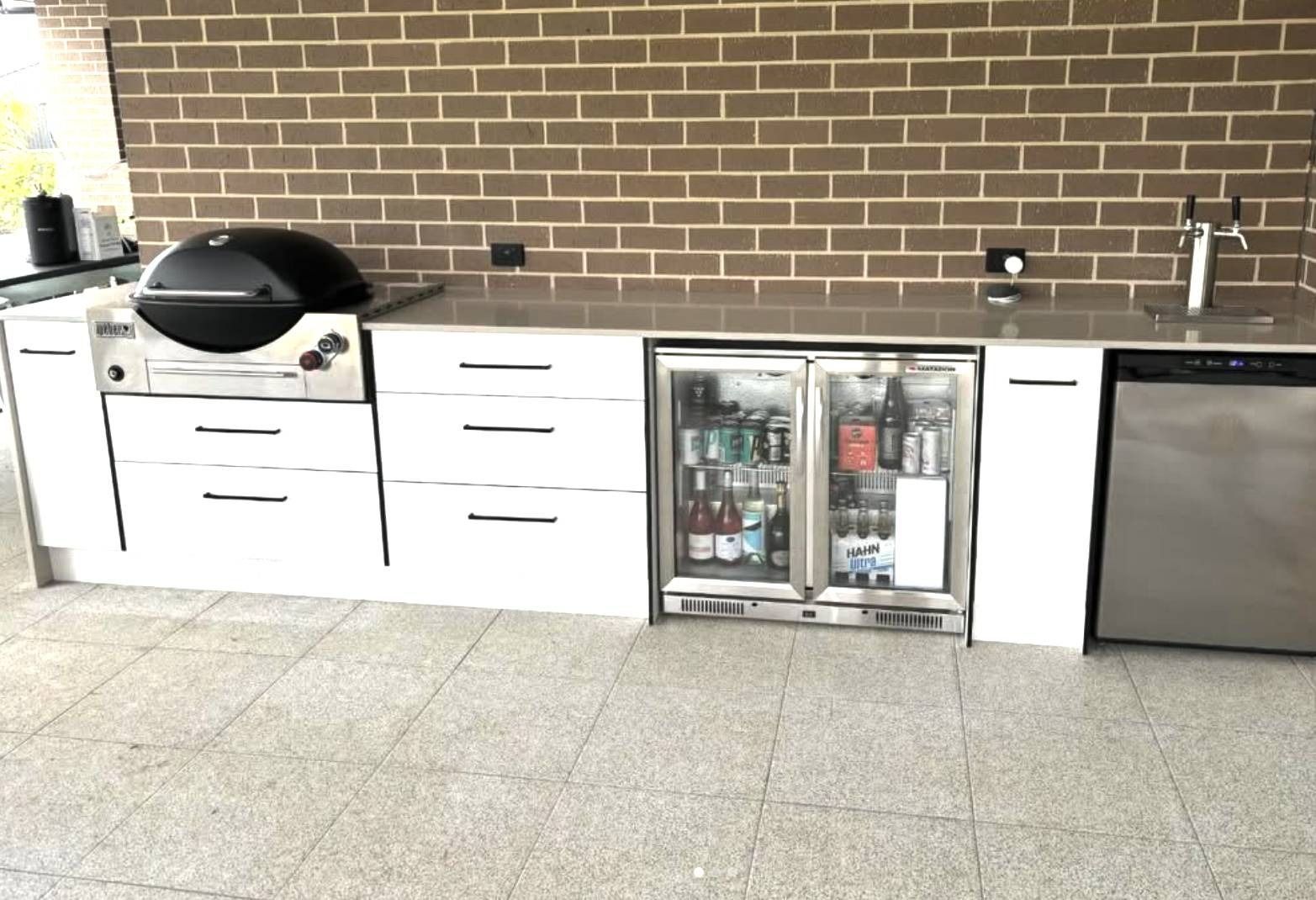 Outdoor kitchen with brick backsplash, white cabinets, grill, mini fridge, and dishwasher