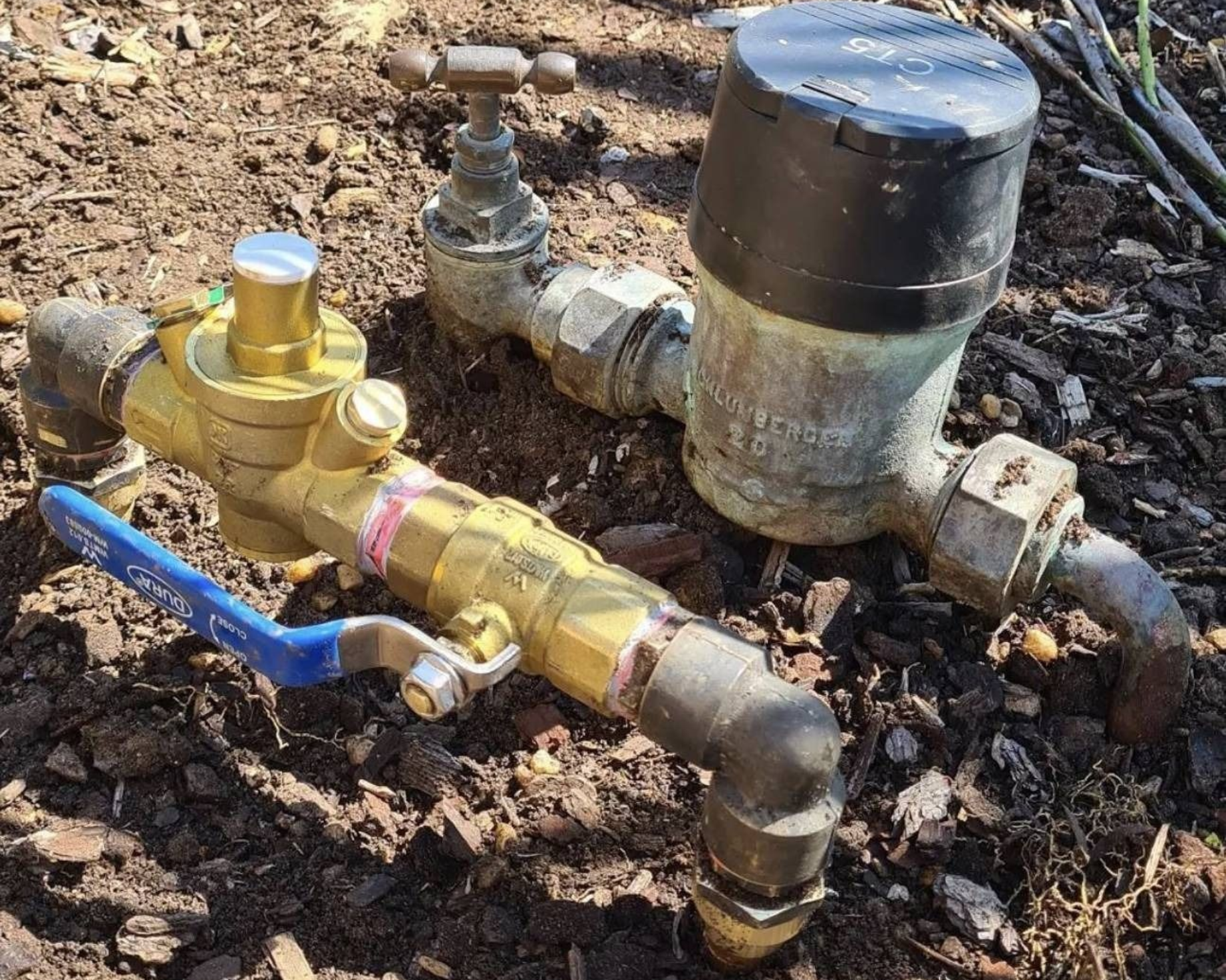 Water meter and brass valve assembly with blue handle connected to pipes outdoors on dirt ground