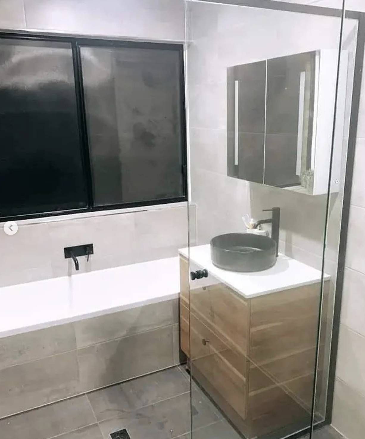 Modern bathroom with gray tile, black-framed window, bathtub, and wood vanity with round sink and mirror.