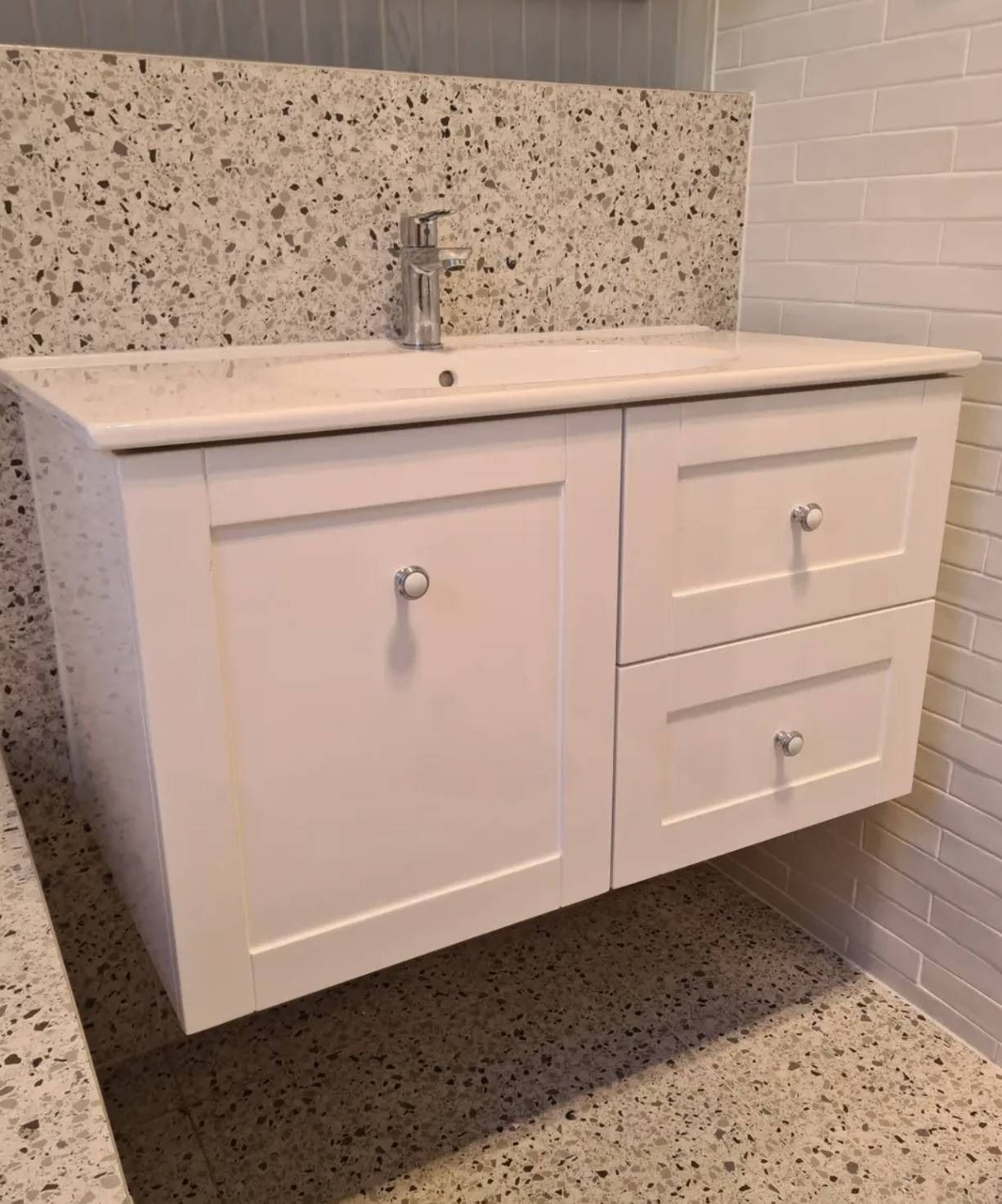 Wall-mounted white bathroom vanity with sink and drawers against speckled and white tiled walls.