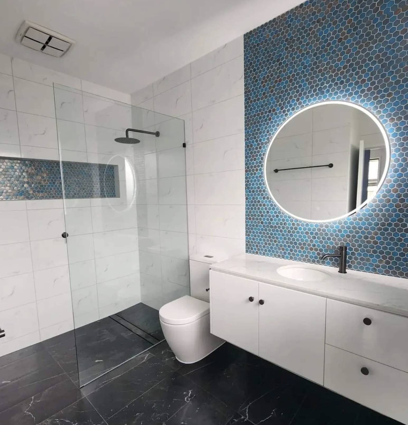 Modern bathroom with blue mosaic accent wall, round lit mirror, white vanity, and glass shower.