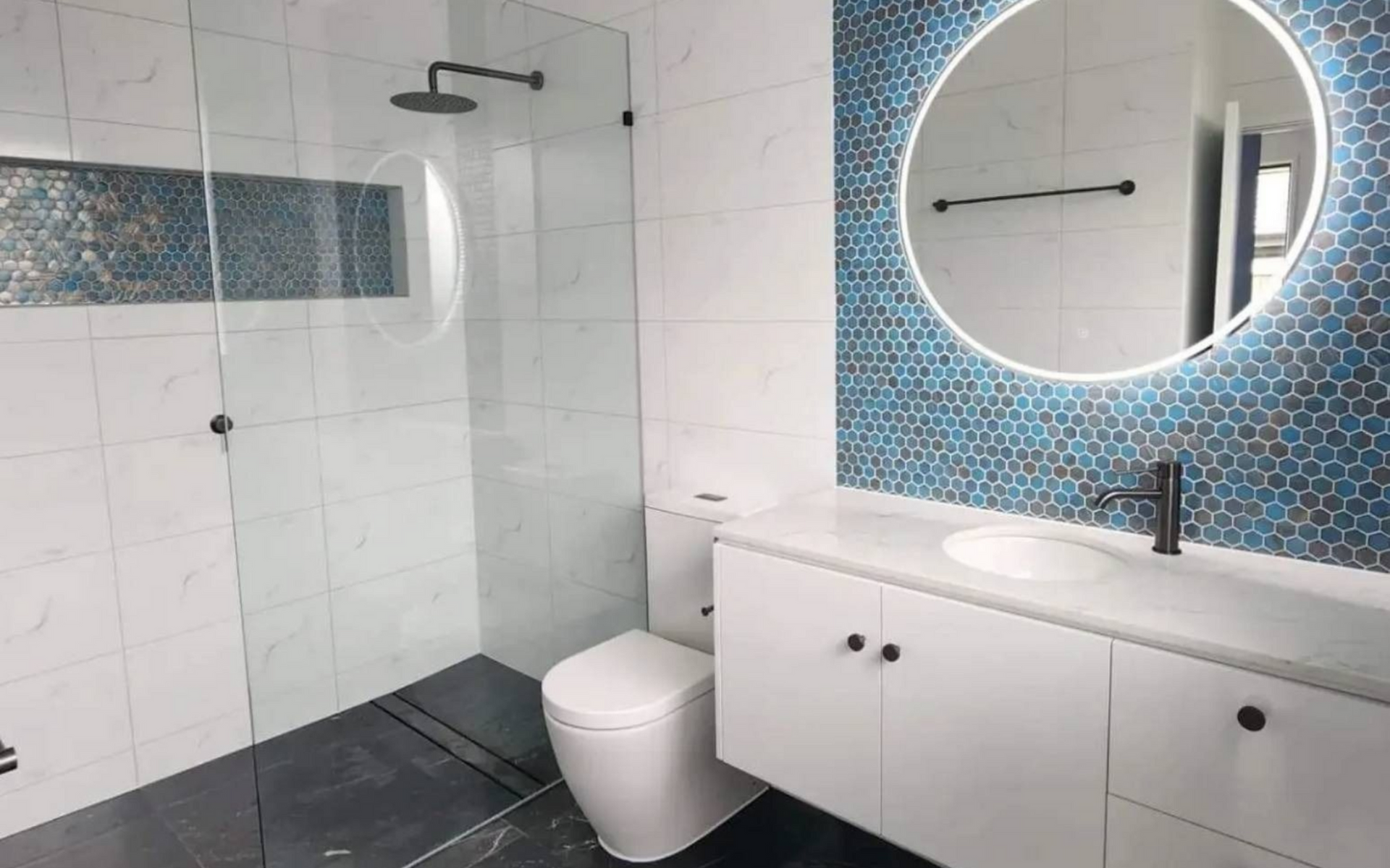 Modern bathroom with glass shower, wall-mounted toilet, white vanity, and round illuminated mirror