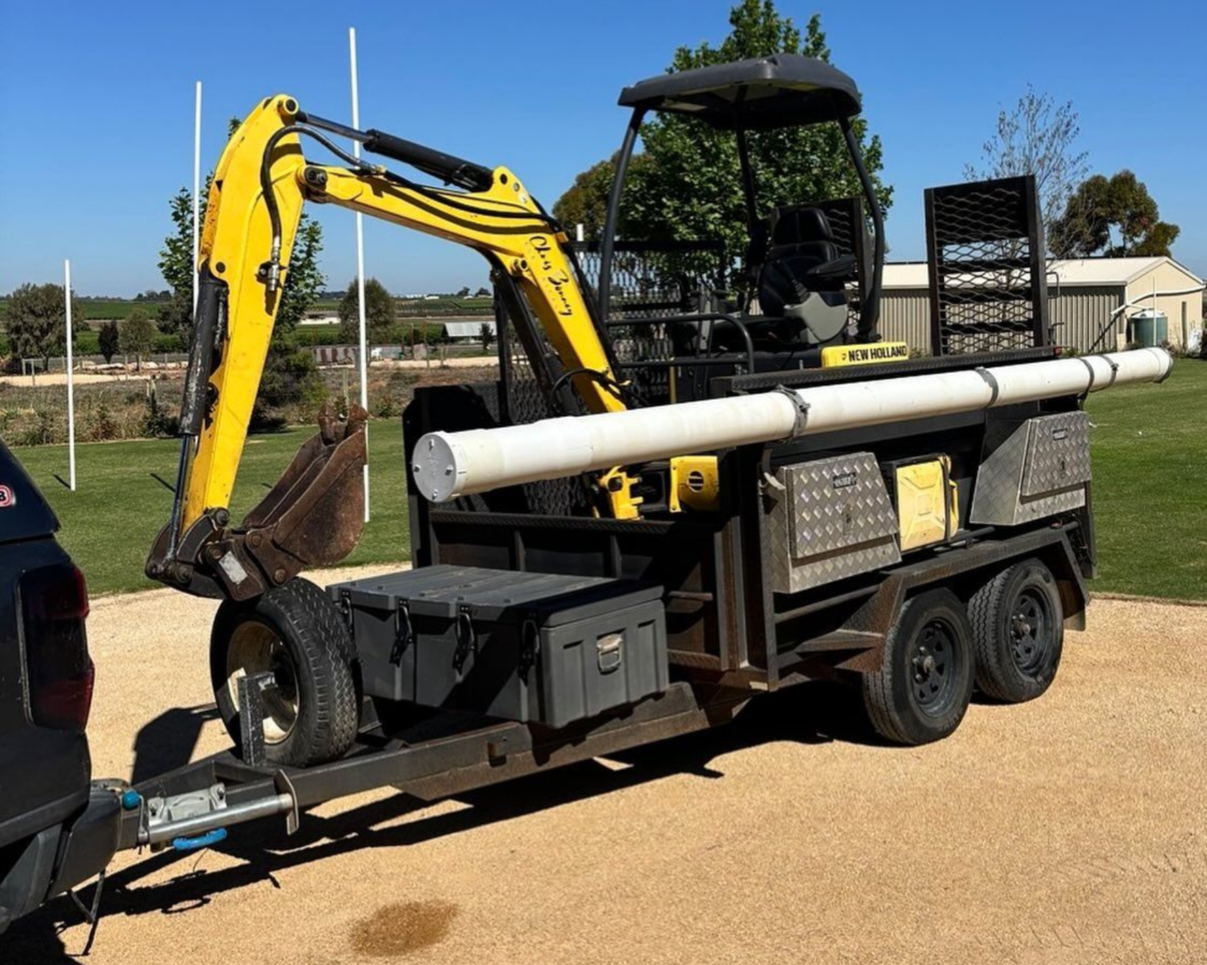Yellow excavator on a flatbed trailer with white pipes strapped to it, parked outdoors under a clear sky