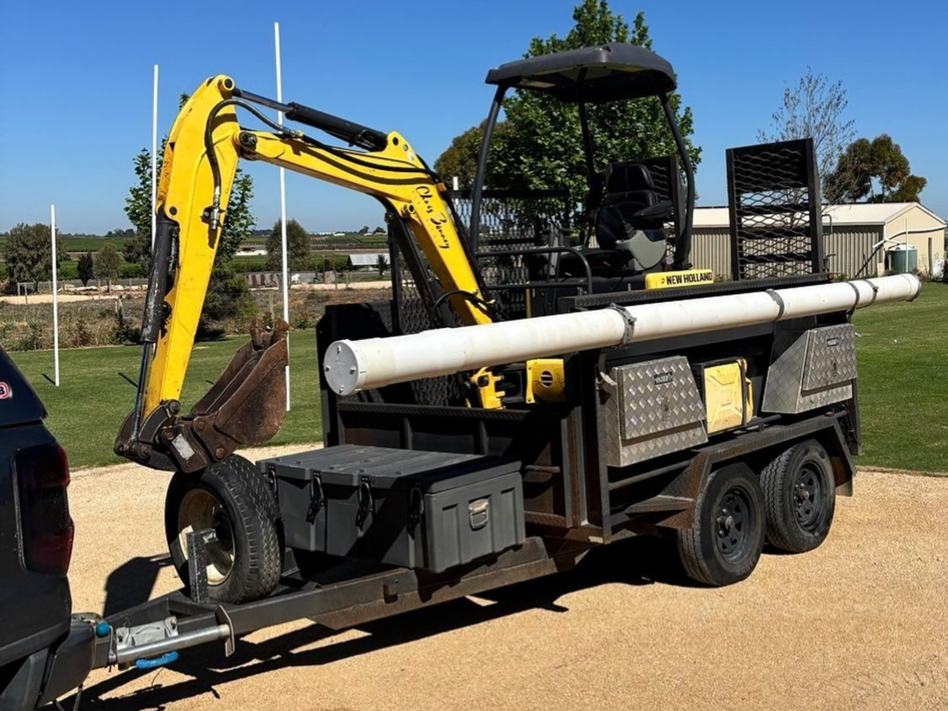 Yellow excavator on a trailer with large white pipes, parked outdoors under a clear sky.