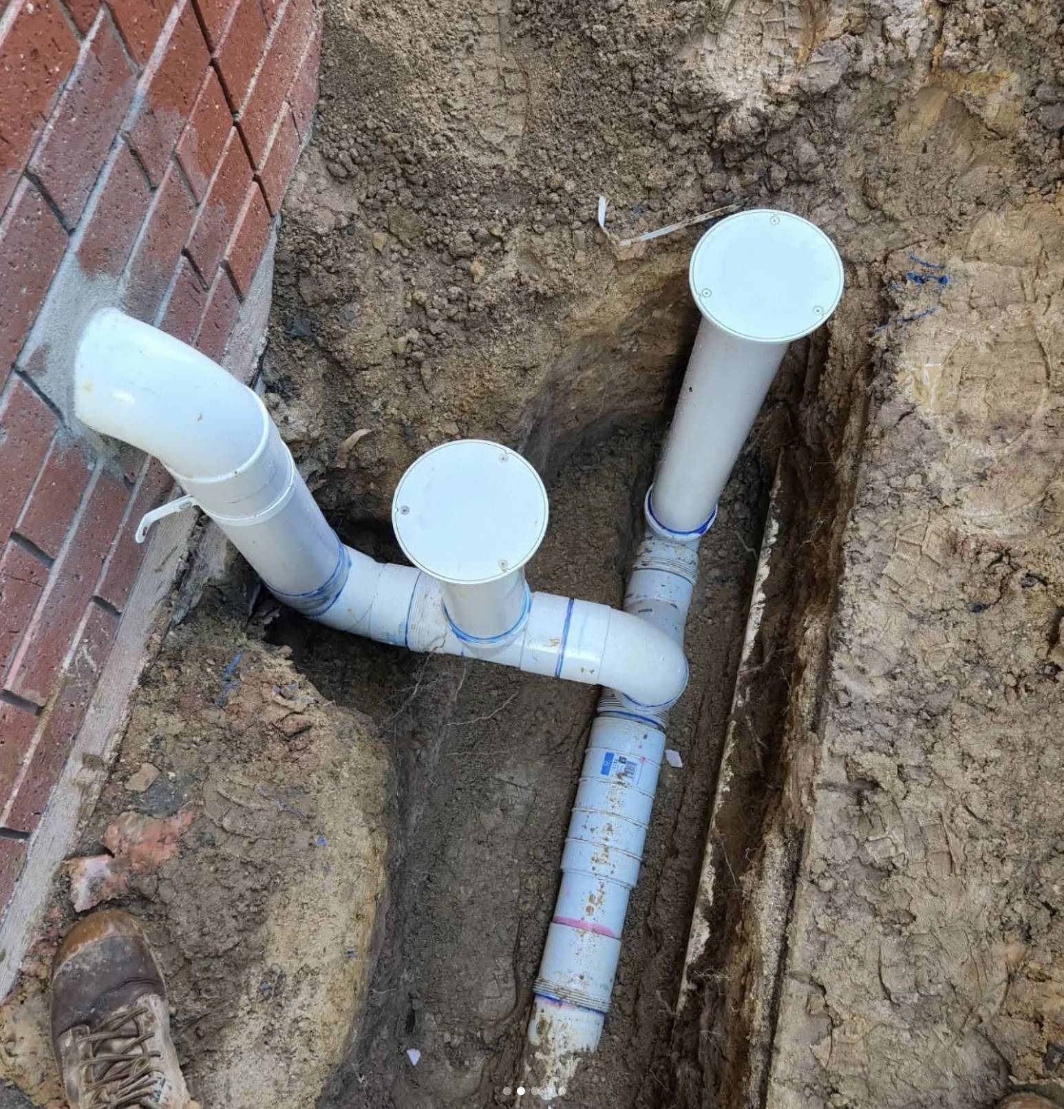 Exposed white PVC pipes and caps in a dirt trench beside a brick wall.