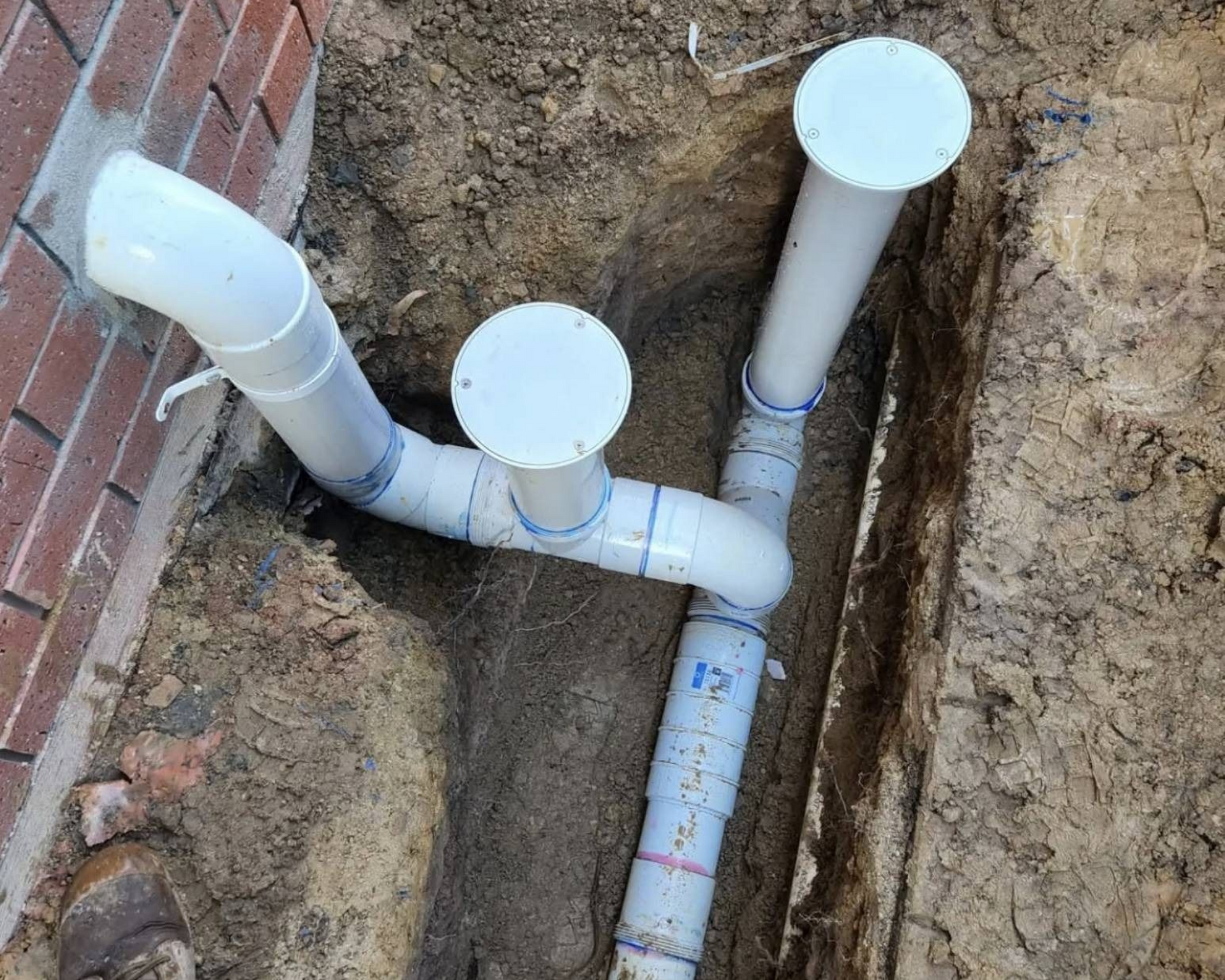 White PVC plumbing pipes in a dirt trench beside a brick wall.