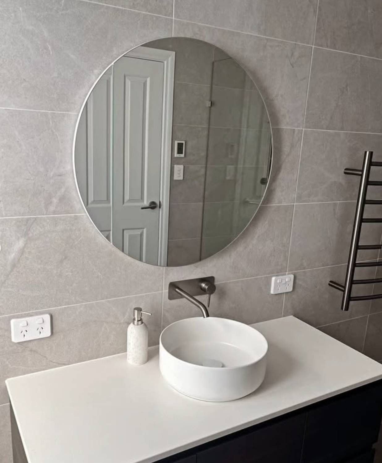 Modern bathroom vanity with round mirror, white vessel sink, and gray tiled wall.