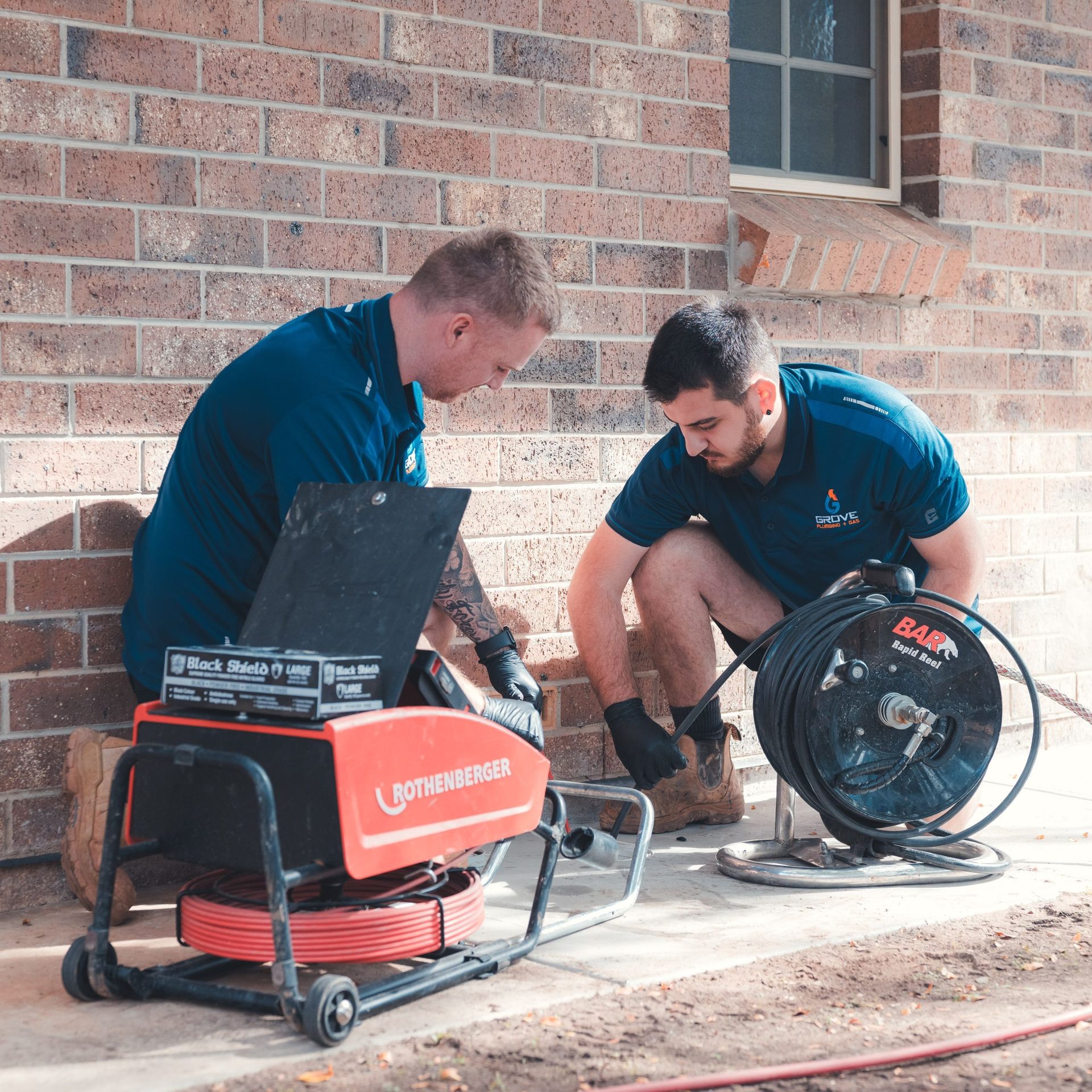 Two workers inspect a red cable machine beside a brick wall outside.