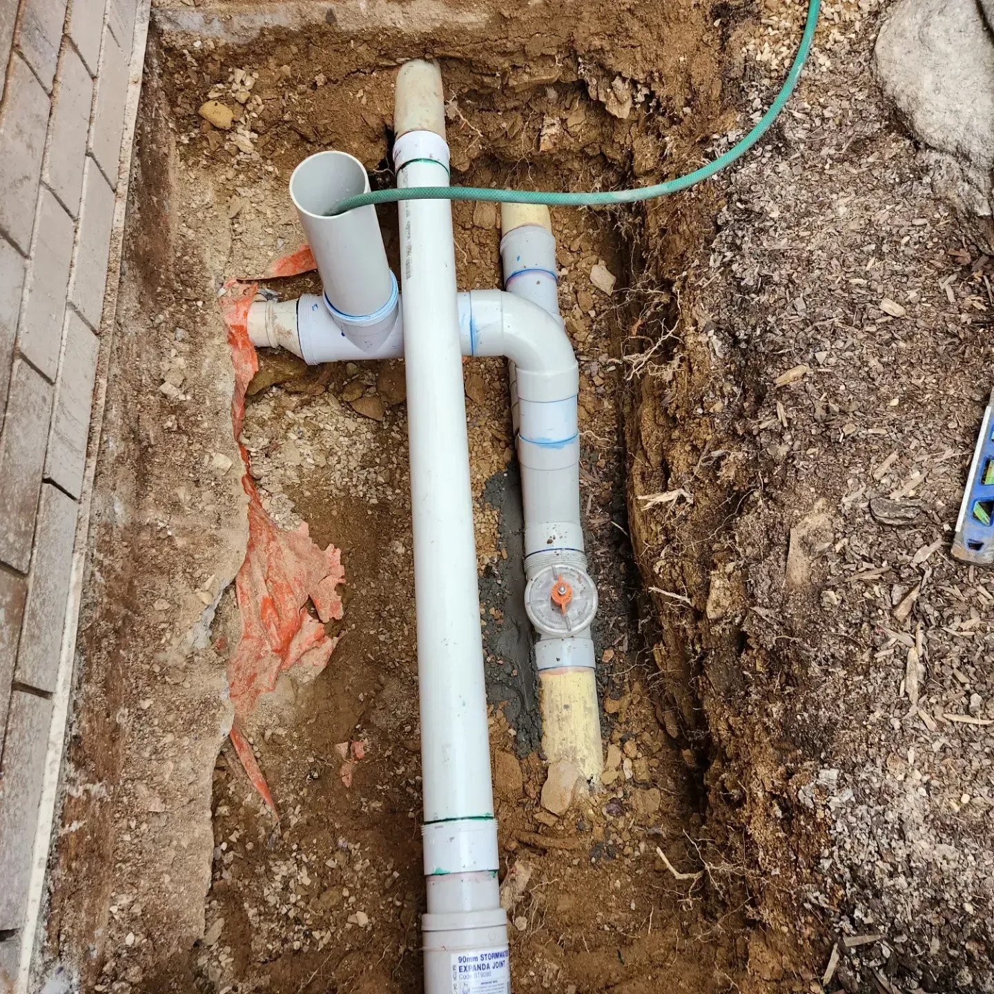 PVC pipes in a dug trench beside a brick wall, with a green hose at the top.