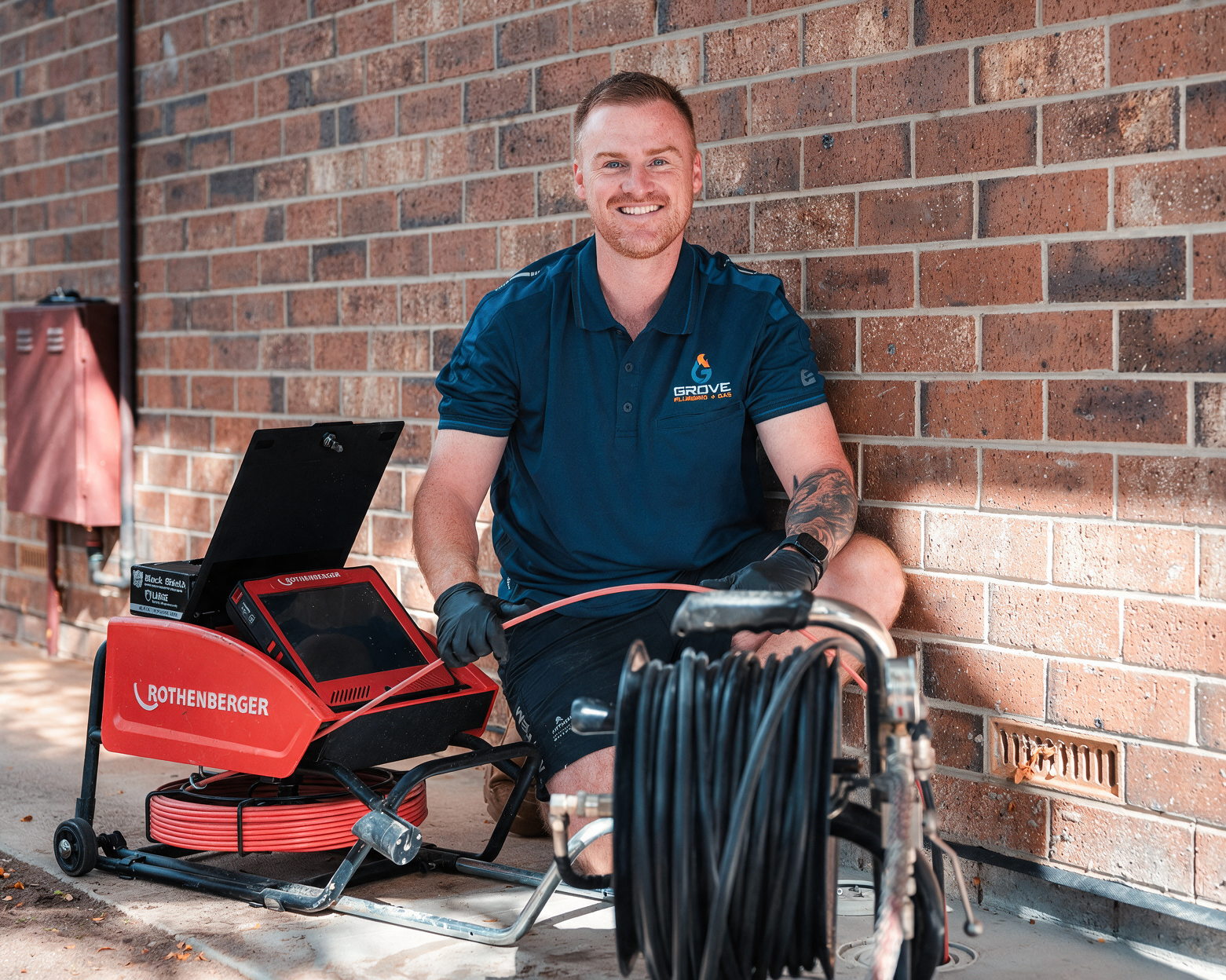 Smiling technician kneels by red drain-cleaning machine and coiled hose against a brick wall