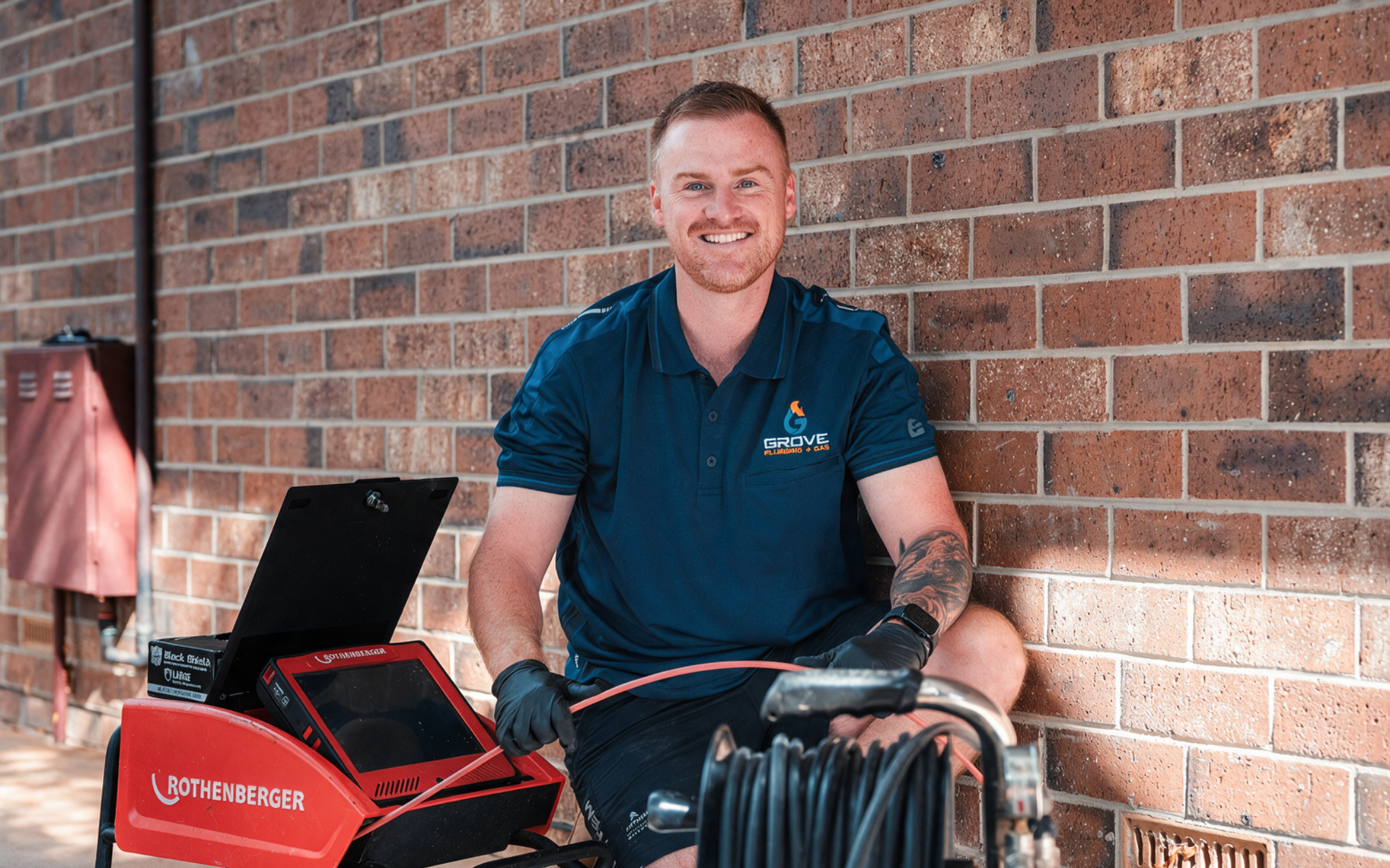 Person in a navy polo stands beside a red grill against a brick wall, smiling outdoors.