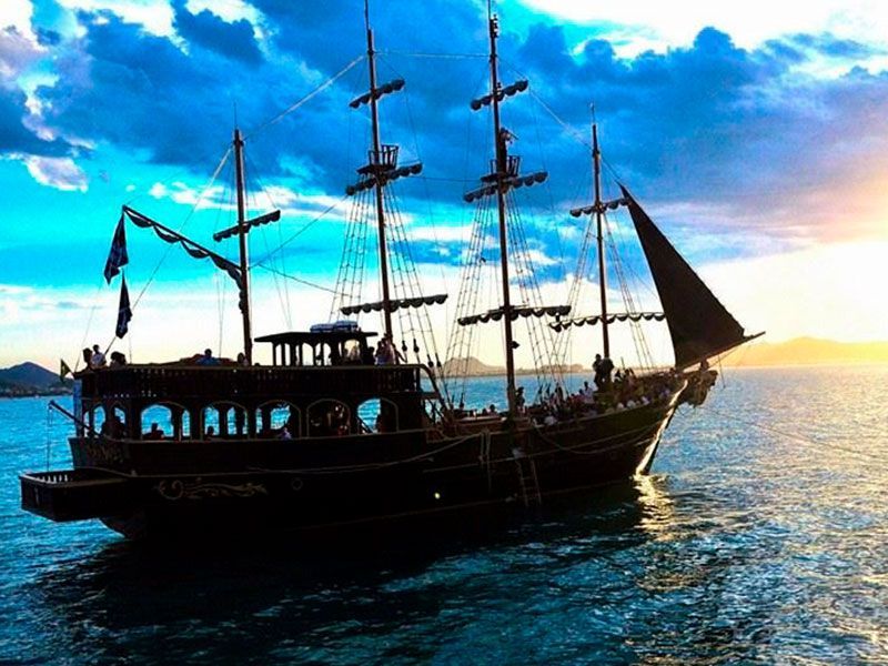 Um grande navio de madeira escura navegando na água com passageiros; céu ao pôr do sol ao fundo.