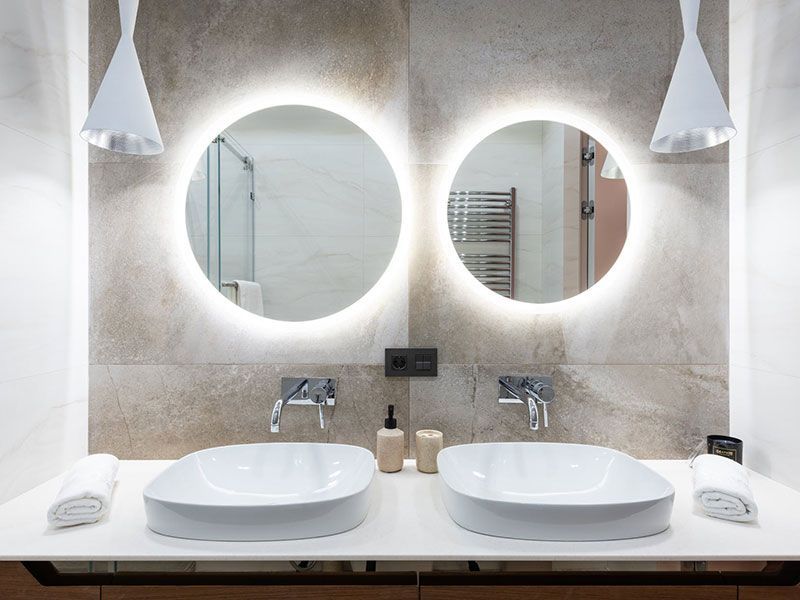Banheiro moderno com duas pias, espelhos redondos com iluminação interna e luminárias pendentes brancas.