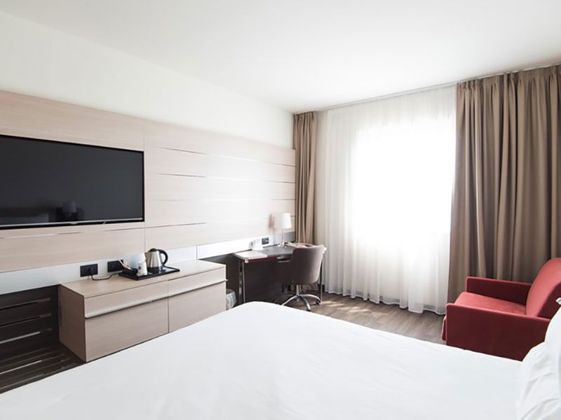 Quarto de hotel com cama, escrivaninha, TV e poltrona vermelha, iluminado por luz natural.