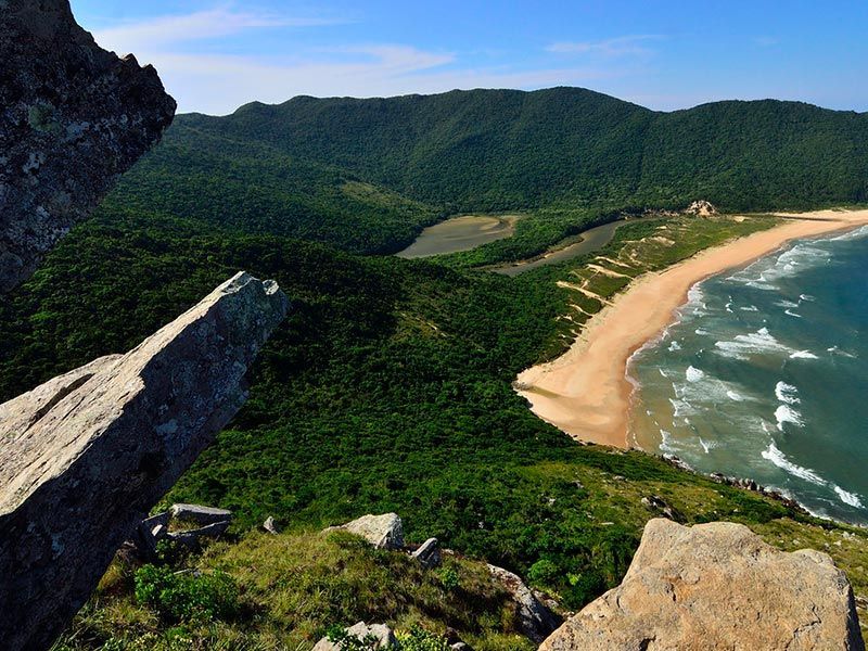 Vista panorâmica de um vale verdejante, praia de areia e oceano sob um céu azul, vista do topo de uma montanha rochosa.
