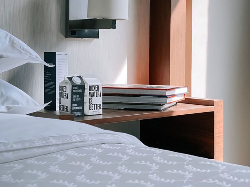 Mesa de cabeceira com pilha de livros, caixa de água, abajur e roupa de cama branca.