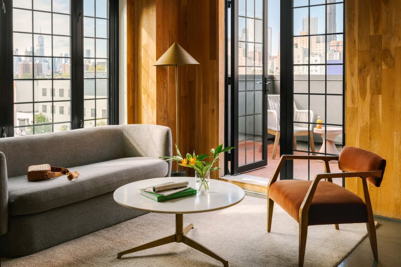 A sunlit living room featuring a gray sofa, white coffee table, and rust-colored chair, with a balcony view of a city.