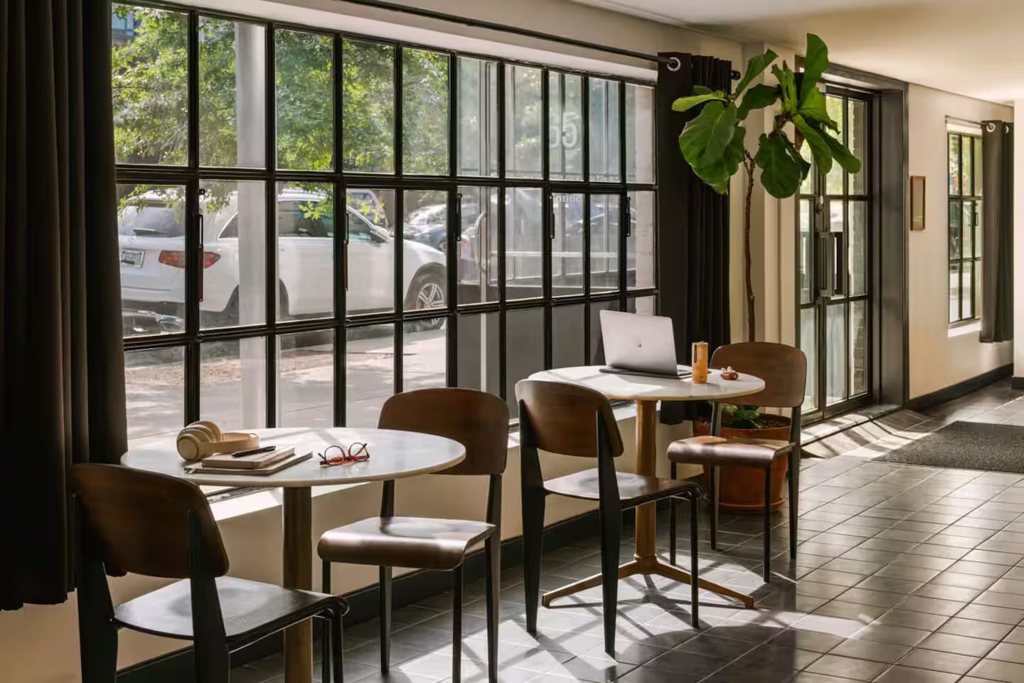 Two round bistro tables with wooden chairs next to a grid-paned window, with a laptop on one table and a plant nearby.