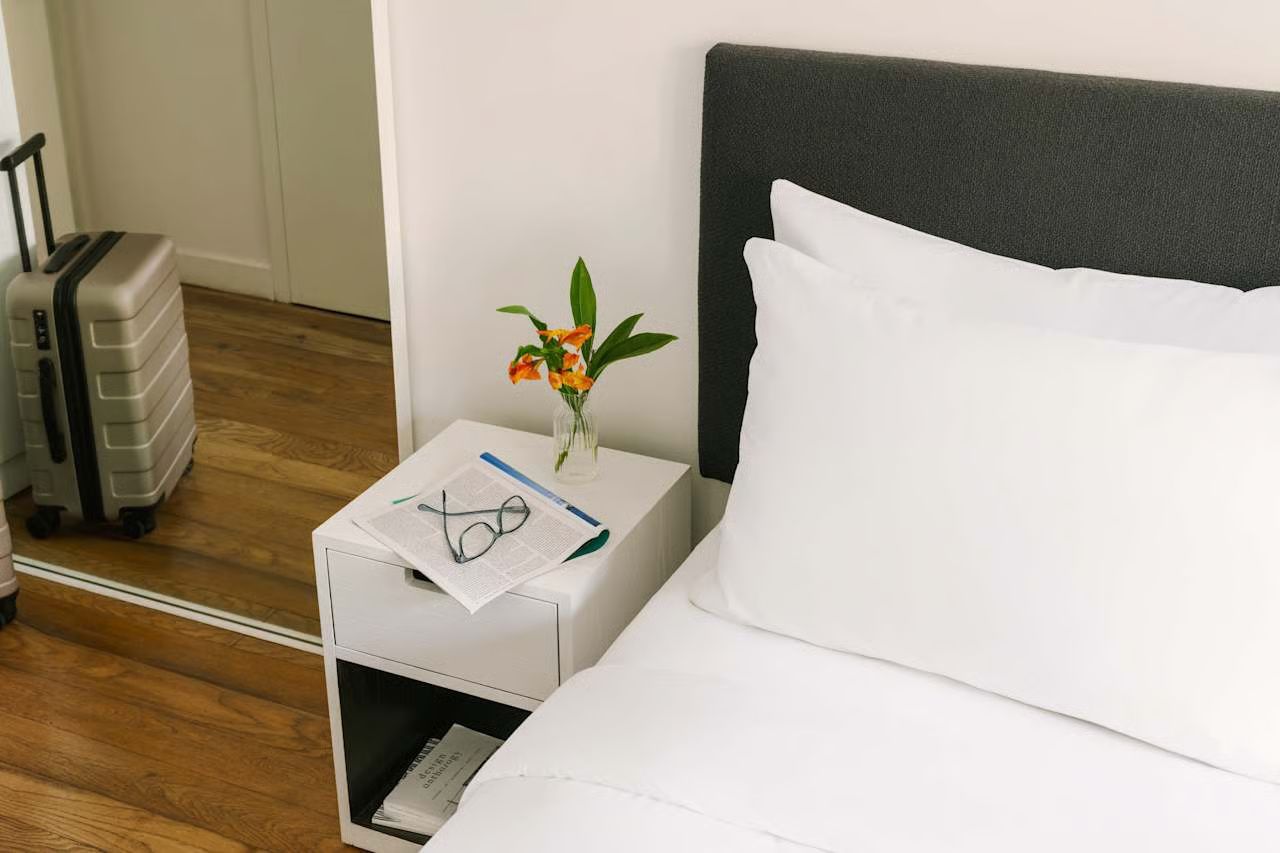 A bedroom scene with a suitcase, white nightstand with a small vase and glasses, and a bed with white bedding.