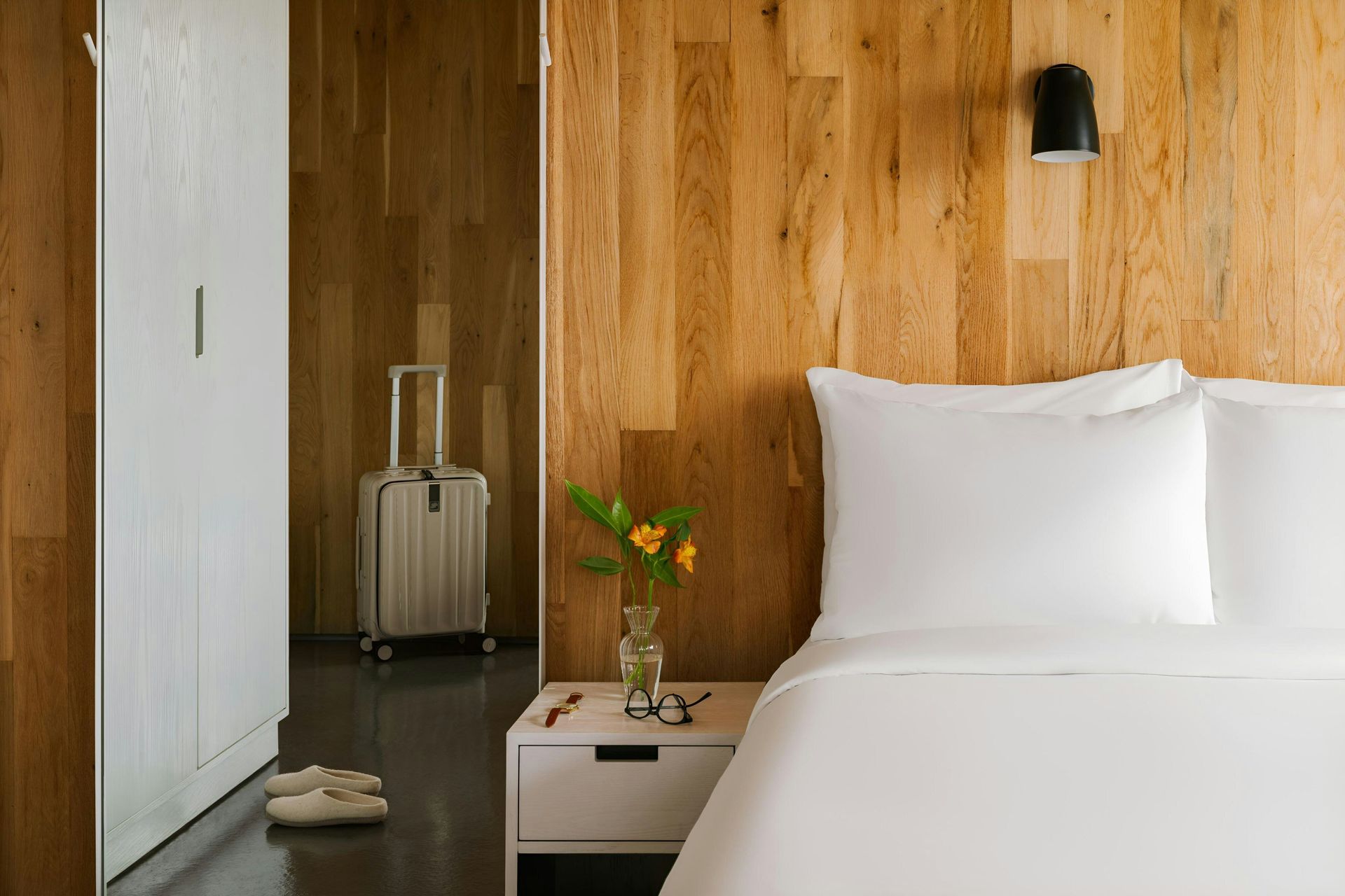 A white bed next to a nightstand with flowers, in a room with wood-paneled walls, a white wardrobe, and a suitcase.