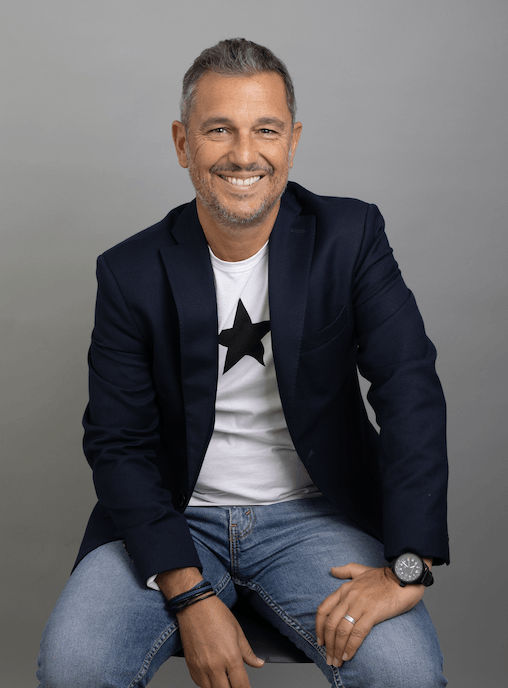 Un homme souriant, vêtu d'un blazer bleu marine et d'un jean, est assis sur un fond gris, les mains posées sur les genoux.
