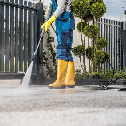 Person in blue overalls and yellow boots pressure washing a concrete surface.